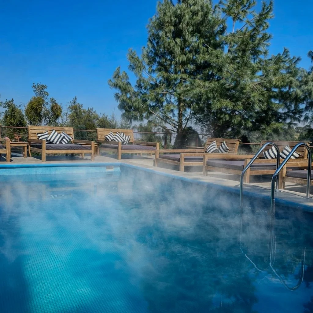 Piscina al aire libre rodeada de sillas de madera con cojines, árboles y cielo azul limpio.