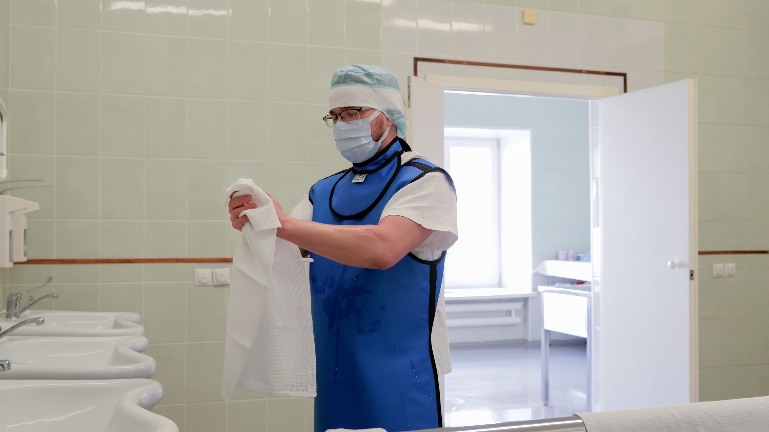 Healthcare worker in a mask and surgical cap wiping down a blue lead apron and thyroid collar between procedures