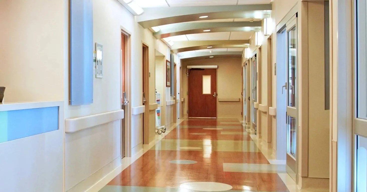 Modern, well-lit hospital corridor with patient room doors and reflective wood flooring.