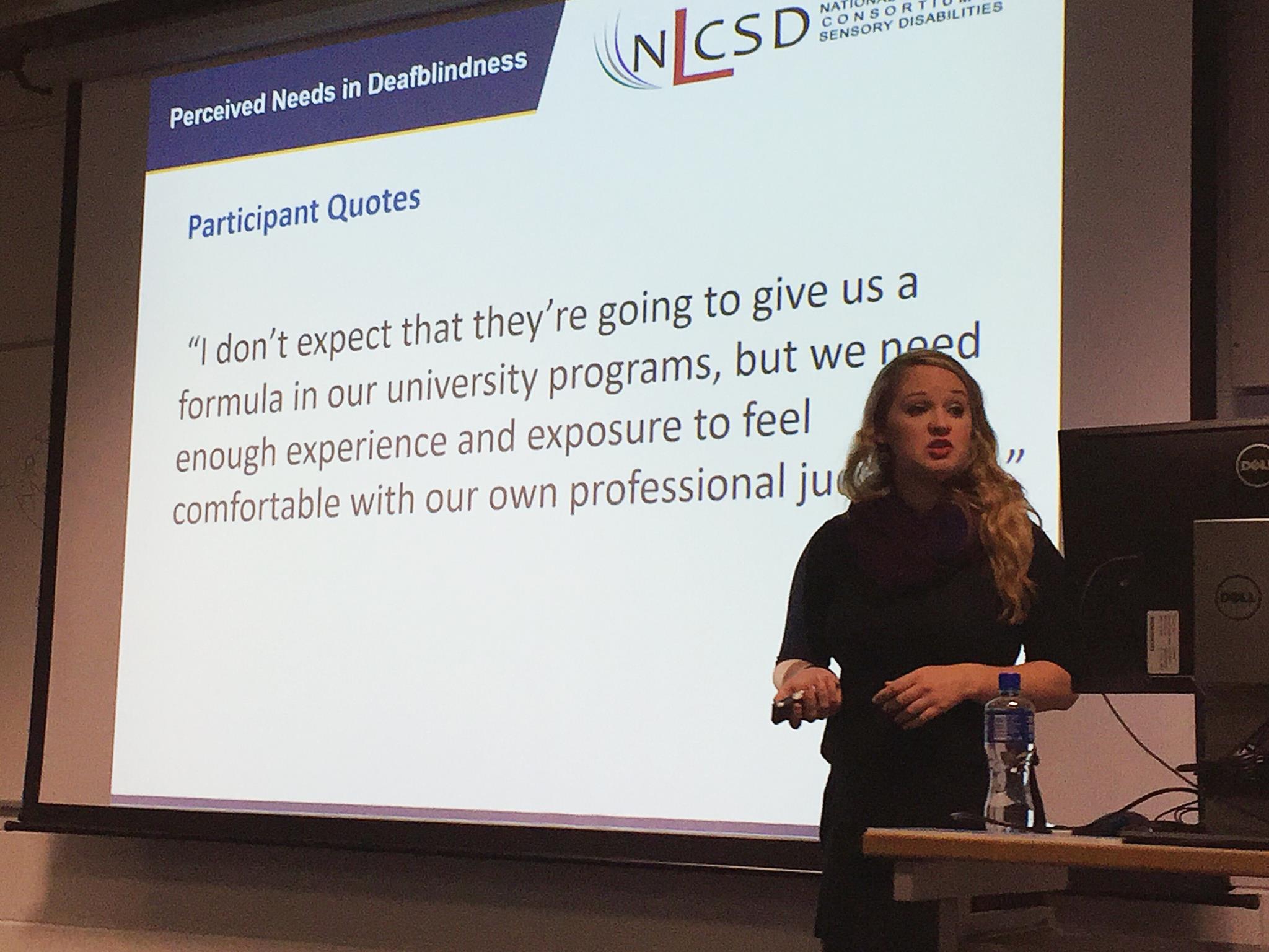 Photo of Tara Brown-Ogilvitanding on a platform in front of a power point projected on a screen behind her.&nbsp;She is wearing a black dress, purple scarf, and black heels while speaking to an audience with a power point clicker in her hand.&nbsp;The power-point encompasses the University of Northern Colorado (UNC) colors in gold and blue. With text that reads, Perceived Needs in Deafblindness: Participant Quote..."I don't expect that they're going to give us a formula in our university programs, but we need enough experience and exposure to feel comfortable with our own professional judgement" with a NLCSD logo in the upper right hand corner.&nbsp;