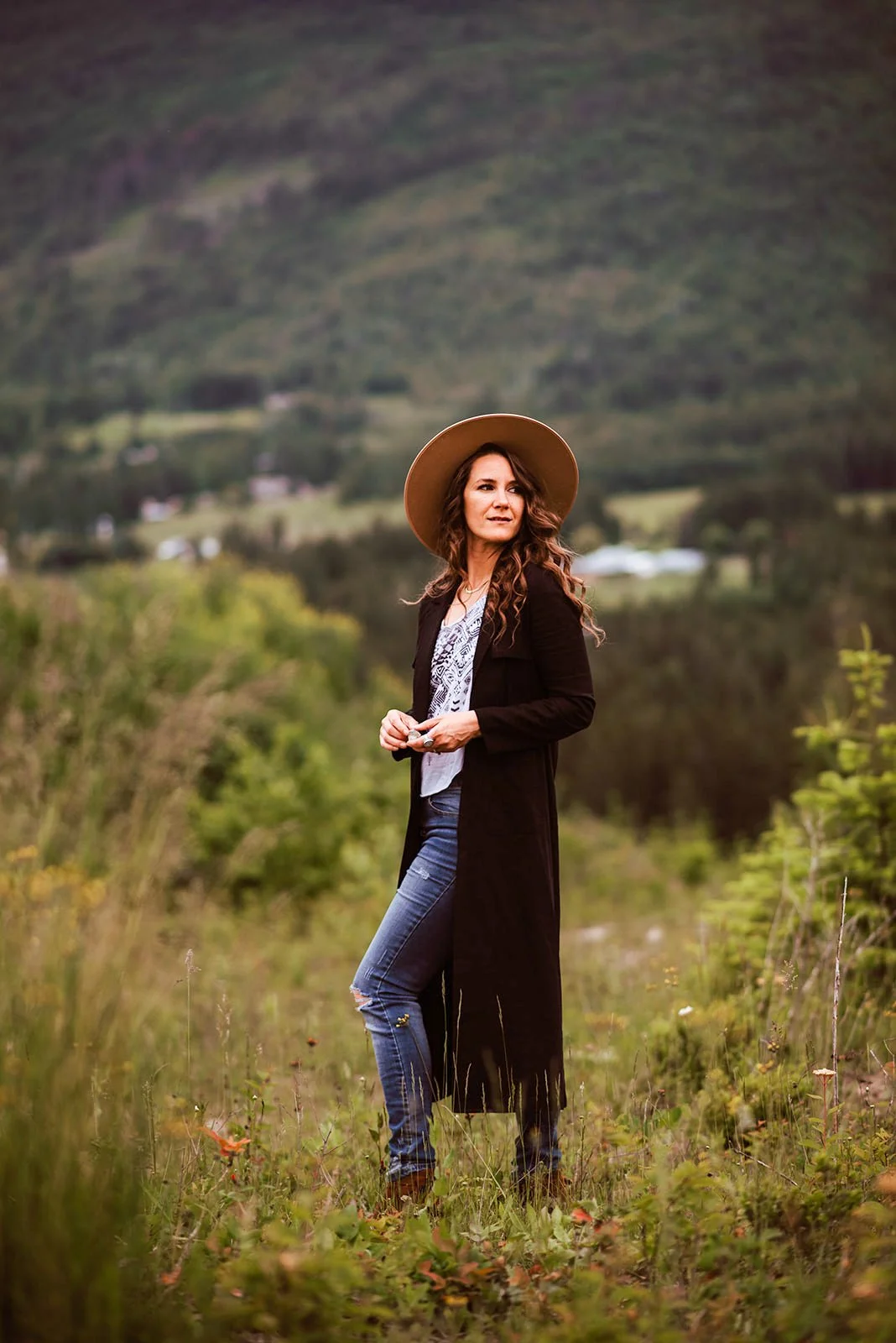 Melissa E Earle aka Mee-woman standing outdoors in a field with mountains and rolling hills in the background. She is wearing a wide-brimmed hat, long black coat, and distressed jeans, holding a camera in her hands and looking off into the distance.