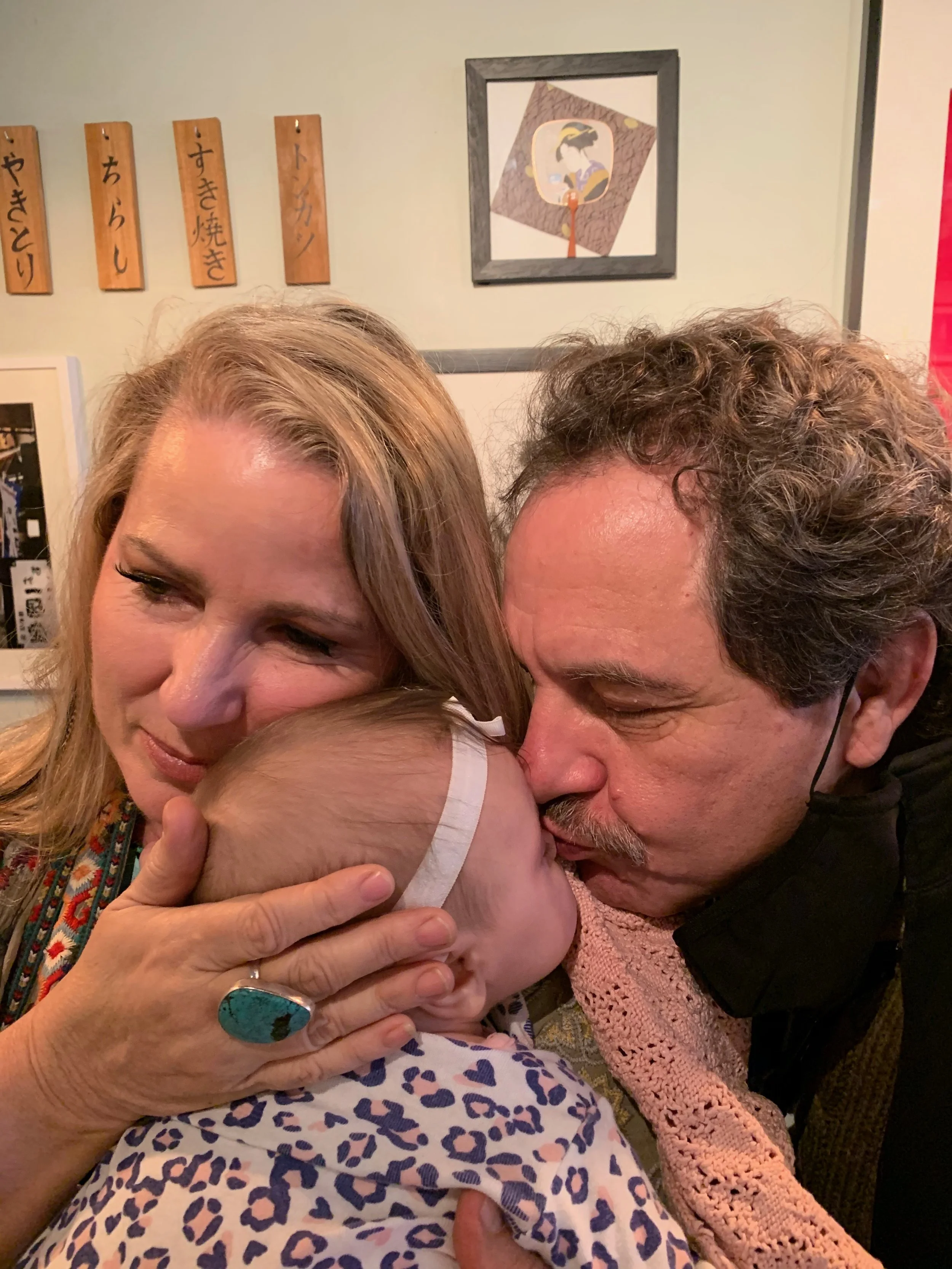 A group of three people, two older adults and a young child, sharing a warm moment of affection and embrace in a cozy indoor setting. The woman has light hair, the man has curly hair and a mustache, and the child has light skin and is wearing a leopa