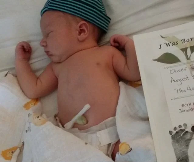 Newborn baby wearing a striped hat, sleeping on a white surface. The baby is shirtless with a muscular build and is positioned next to a birth announcement card.