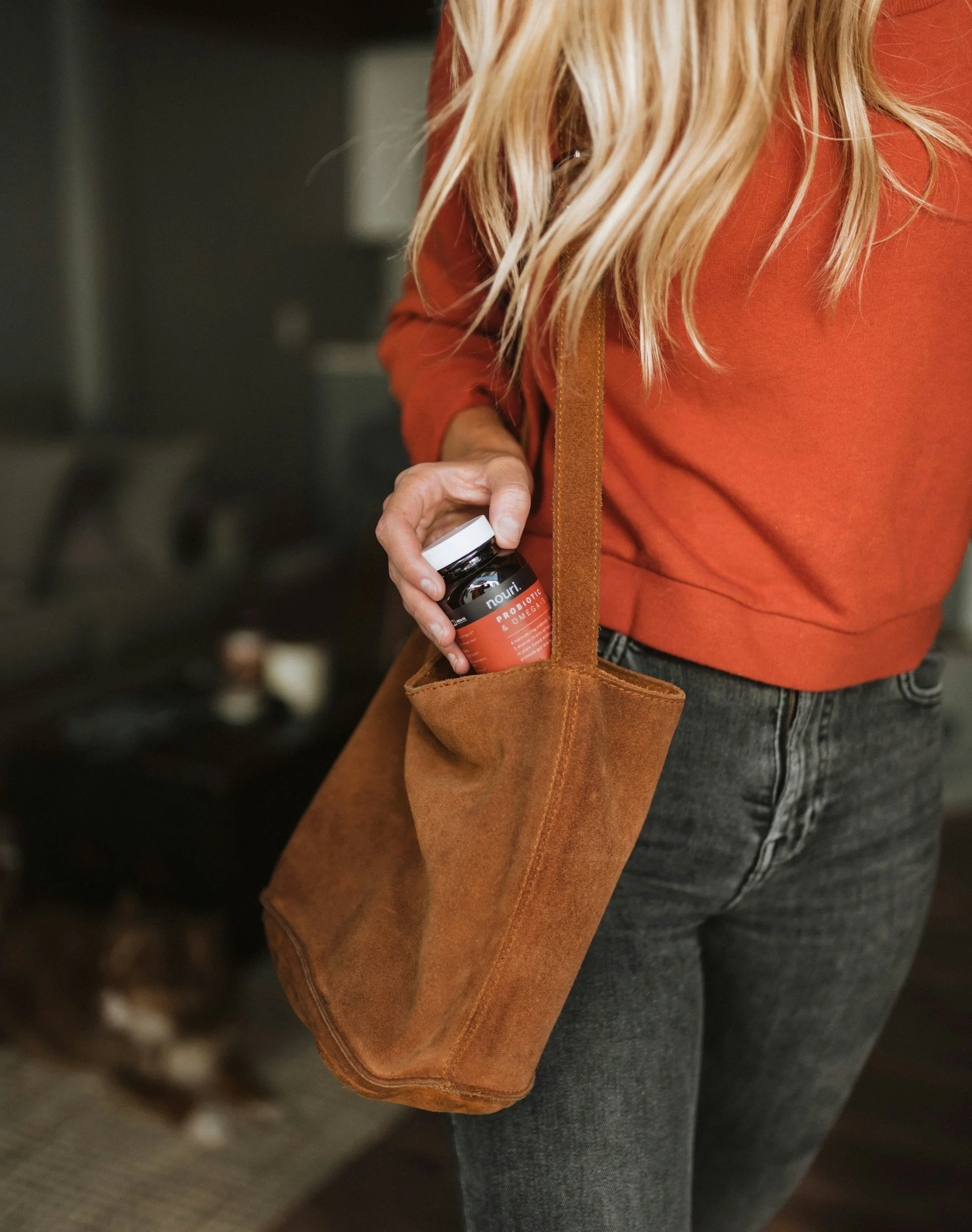 A person in a red/orange long-sleeve shirt placing a bottle of Nourish probiotic & omega supplement into a brown suede tote bag. The person is wearing gray jeans, and the background is blurred.