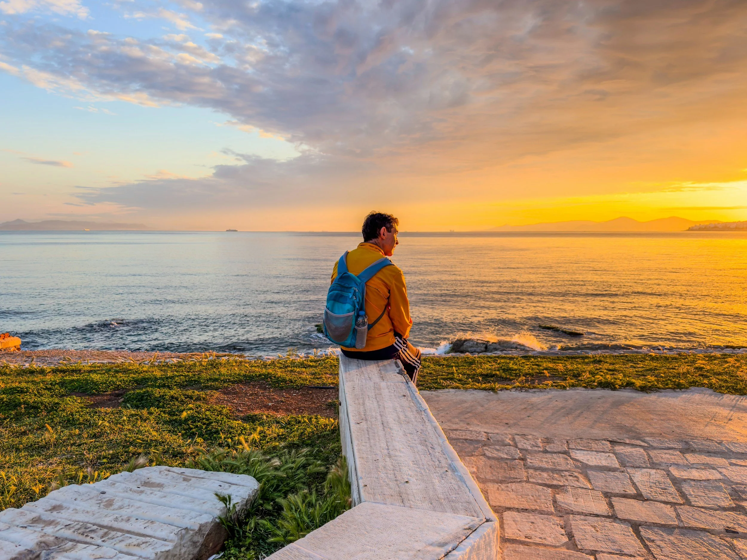 A person with dark hair wearing a yellow jacket and blue backpack sitting on a white bench facing the water during sunset, with sky filled with clouds and distant ships on the horizon.