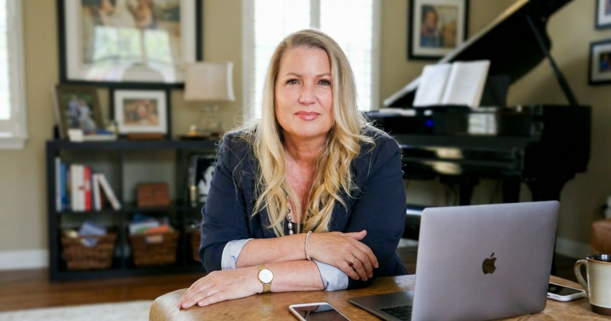 A woman sitting at a desk in a home office, with a laptop, smartphone, and coffee mug, in front of a piano and framed photos on the wall.