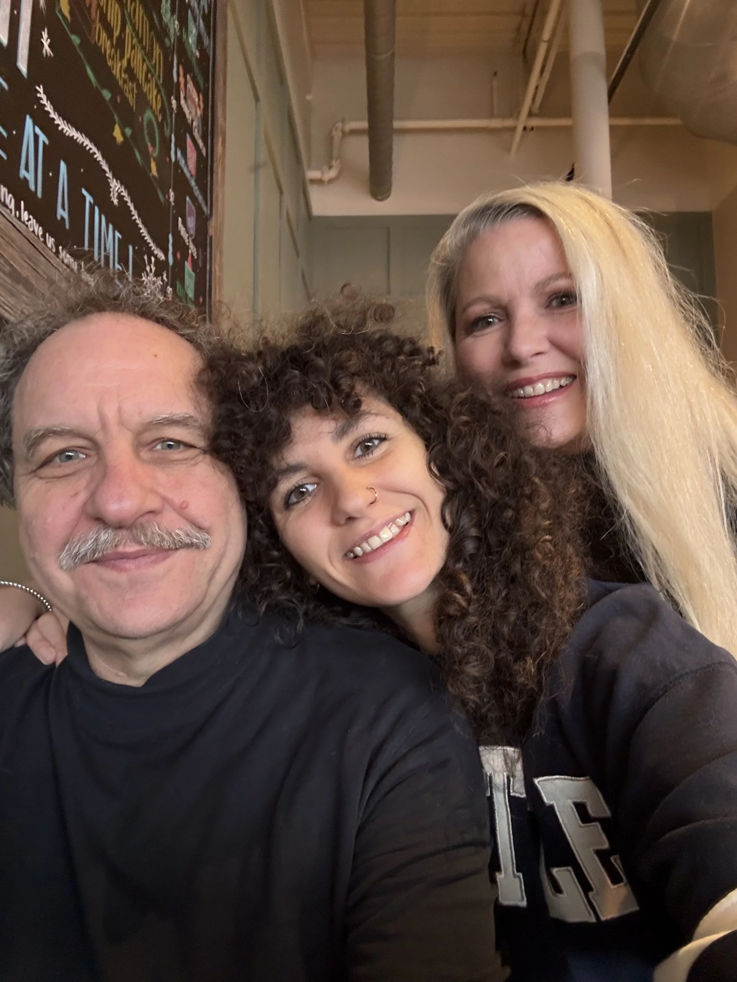 Three people taking a selfie together, smiling, in what appears to be a casual indoor setting with exposed pipes and a blackboard in the background.