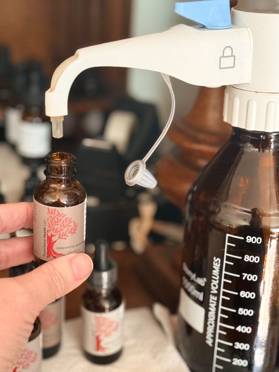 A person holding a small brown bottle labeled 'Supernatural Health LLC' under a white spray bottle used for dispensing liquids, with other bottles in the background.