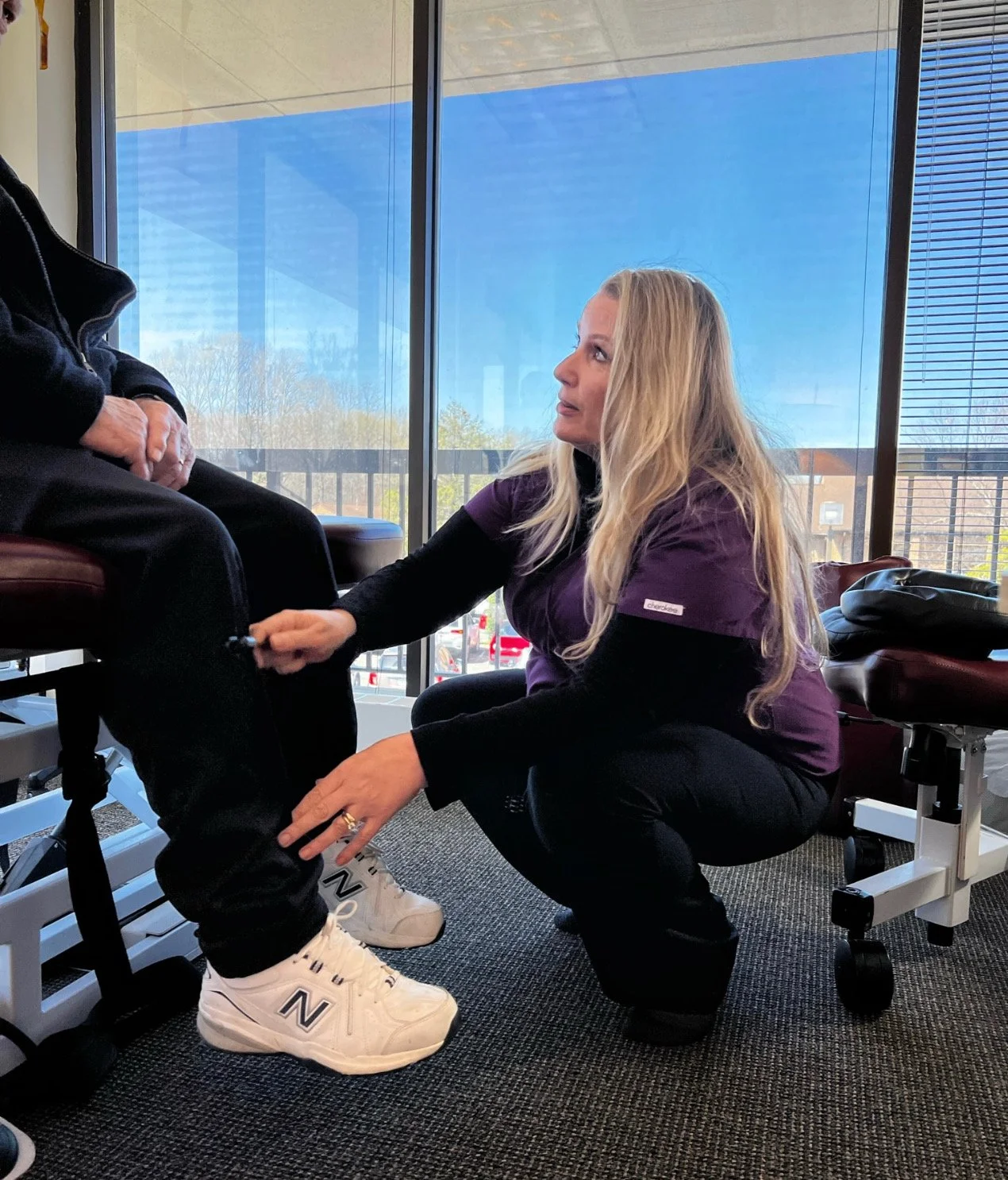 Hospital room with an examination bed, a stool, a red blanket, a bedside lamp, and large windows with blinds showing trees and parked cars outside.