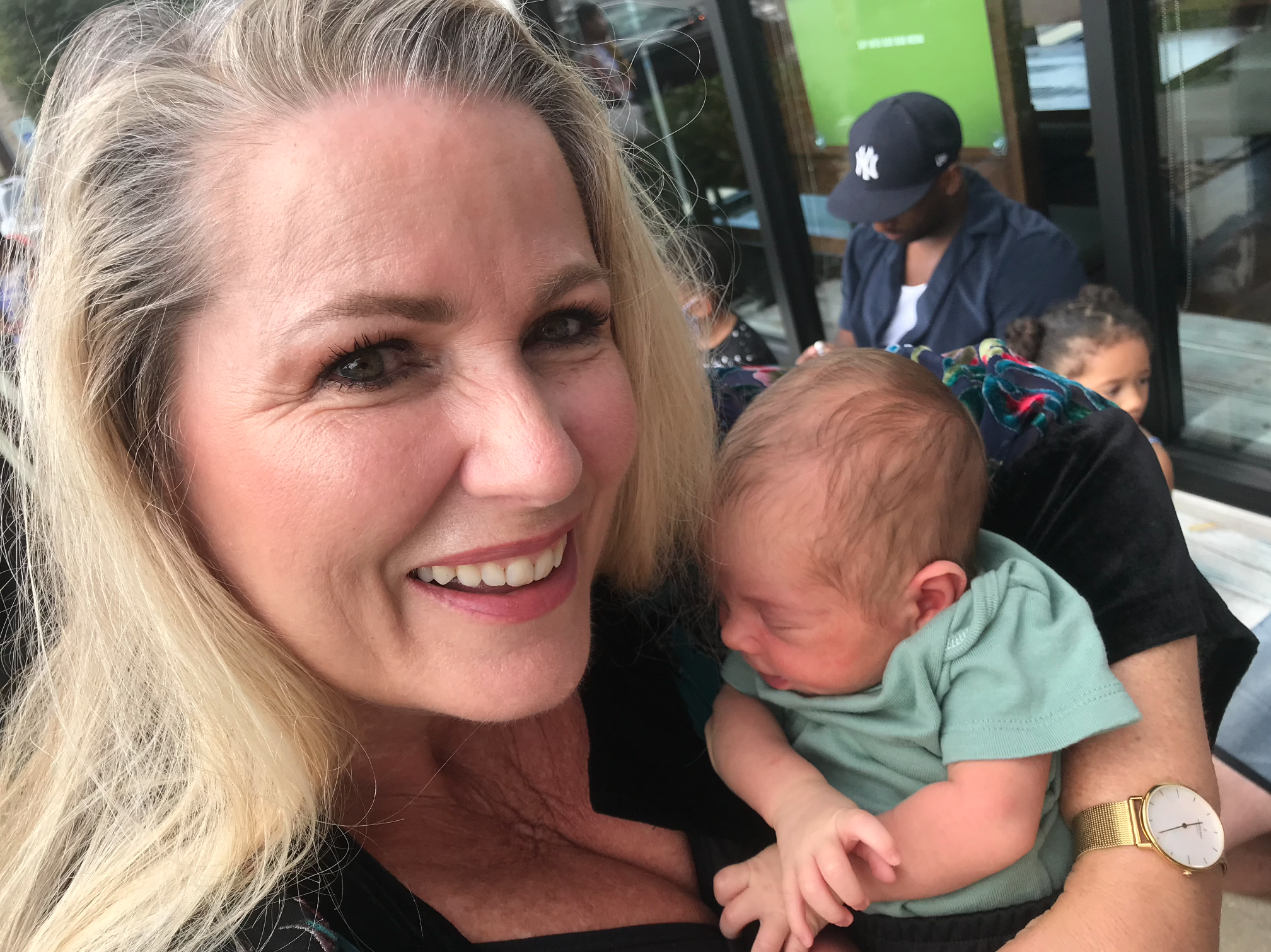 A close-up of a smiling blonde woman holding a baby and standing outdoors with a man in the background wearing a navy baseball cap and glasses, sitting in front of a green sign and glass windows.
