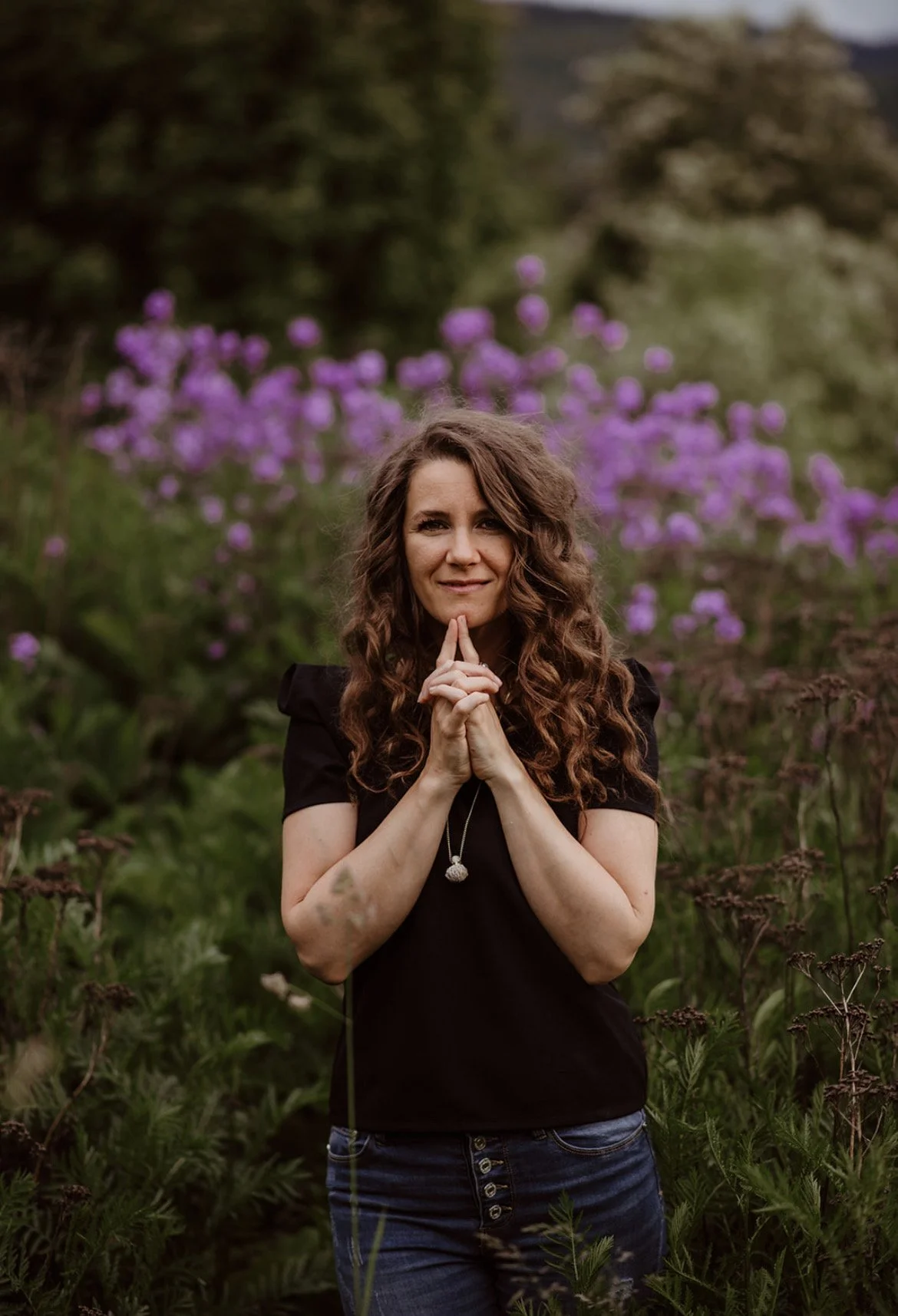 Melissa E Earle aka Mee A woman with long curly brown hair and a black top stands outdoors in a garden with purple flowers in the background, holding her hands together in a prayer-like pose and smiling gently.