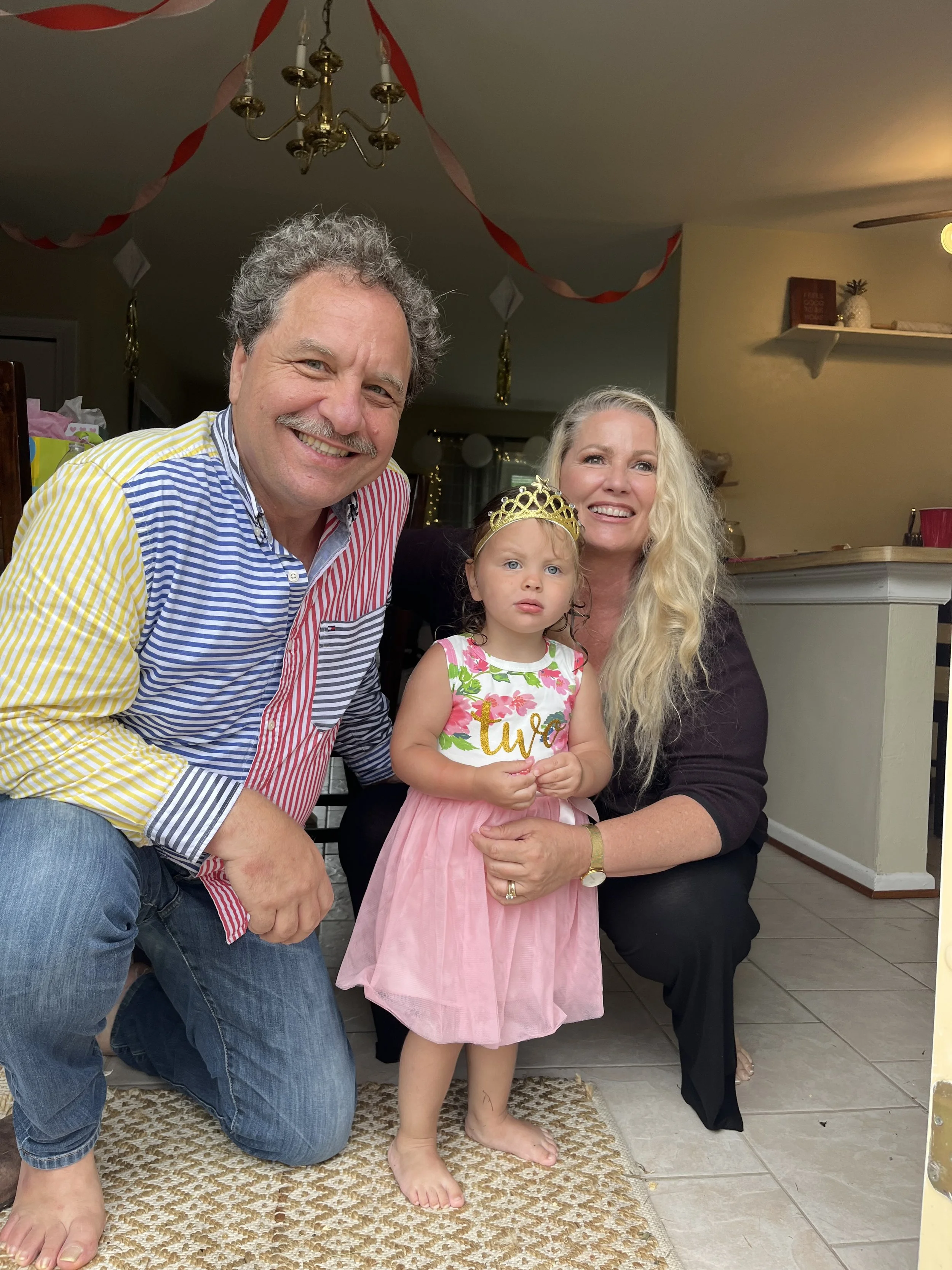 A family of three celebrating at a birthday party indoors, with decorations and a chandelier overhead. The man is kneeling on a rug, smiling, wearing a colorful striped shirt, while a woman and a young girl in a pink dress and a birthday tiara pose w