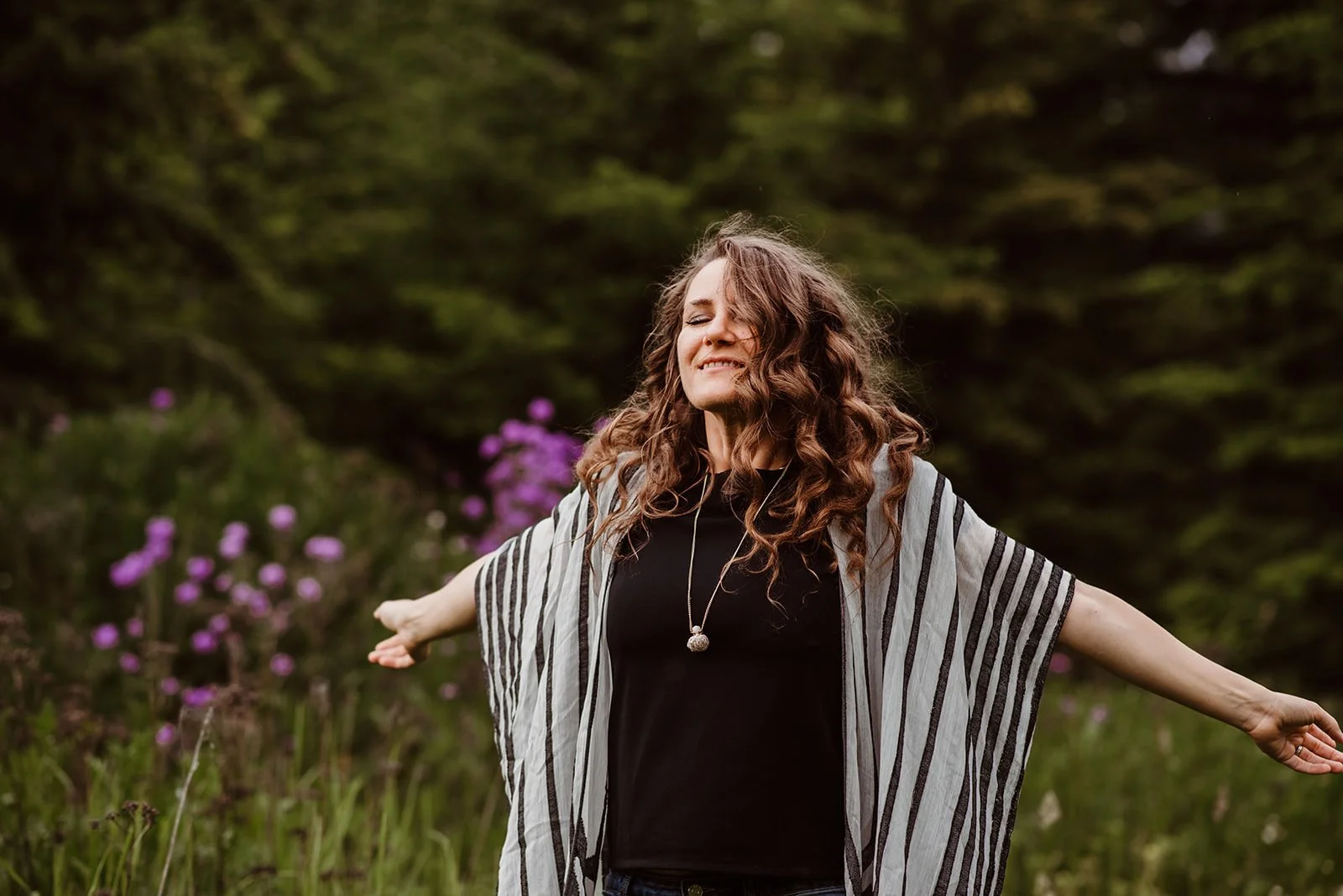 A woman with curly brown hair stands outdoors with arms outstretched, eyes closed, smiling, wearing a black shirt and a striped shawl, with purple flowers and green trees in the background.