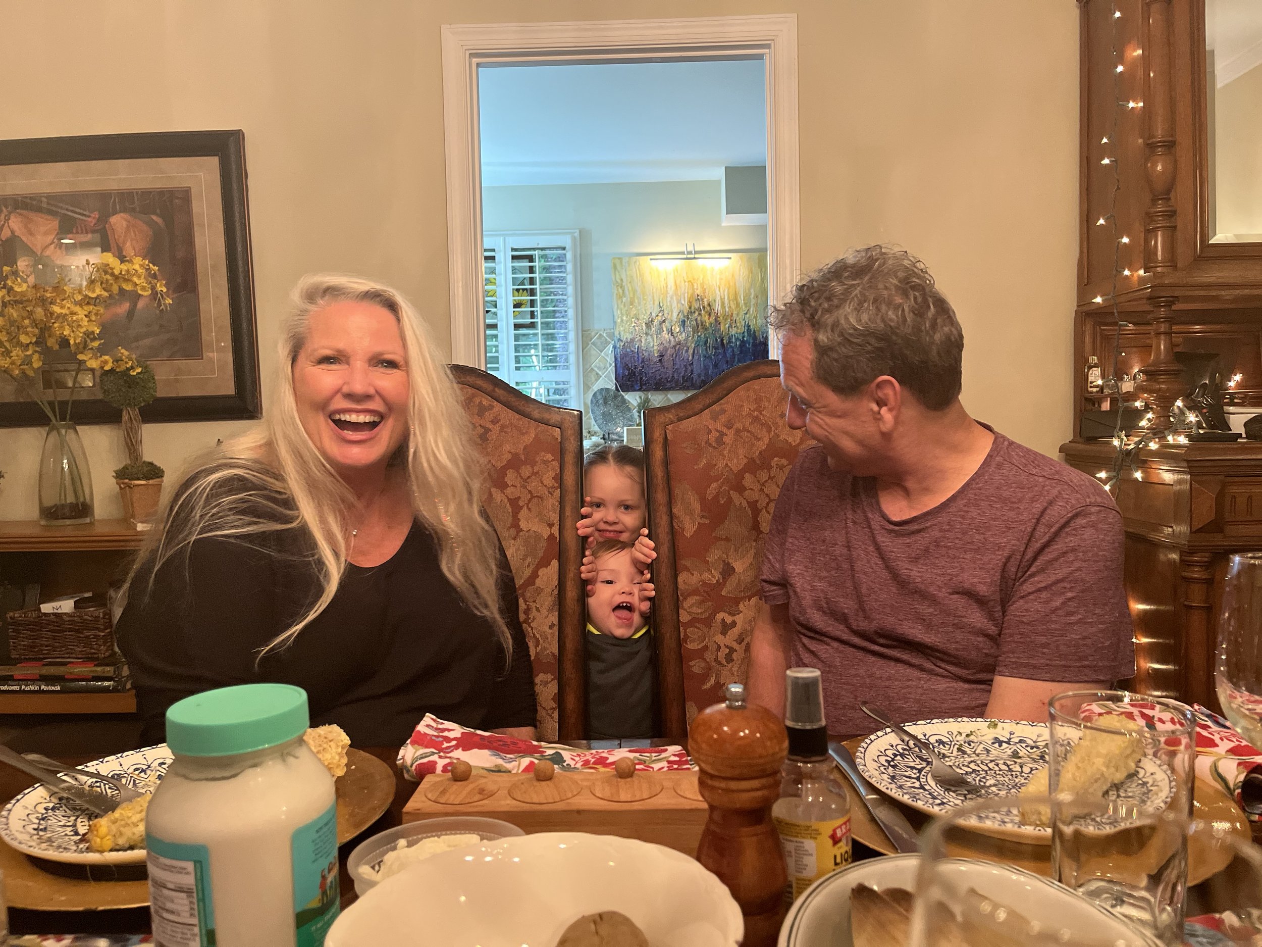 People gathered at a dining table enjoying a celebration, with a woman smiling, a man looking at children, and two children peeking through a decorative photo frame.
