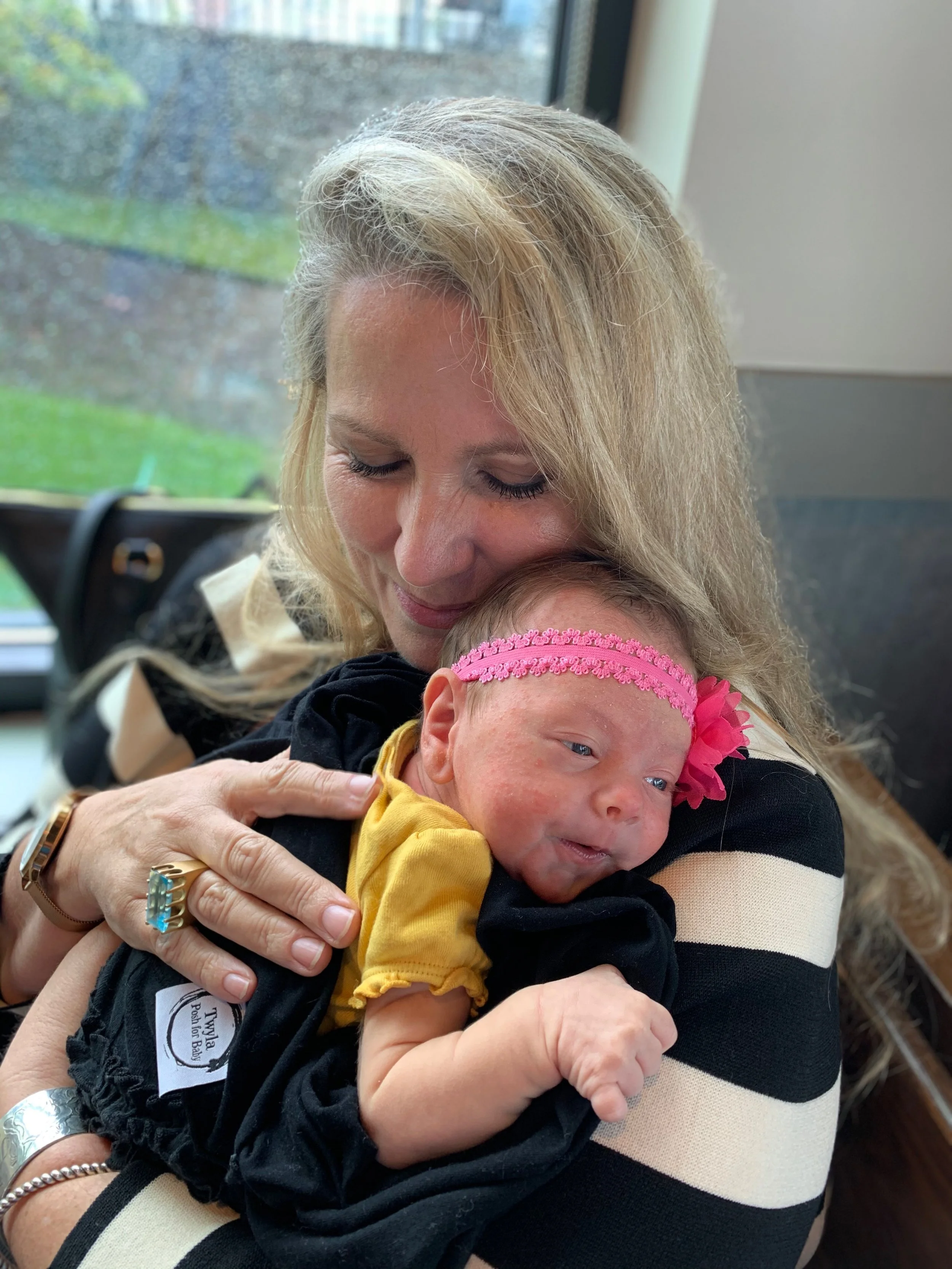 A woman with blonde hair hugging a newborn baby with a pink headband and a yellow outfit.