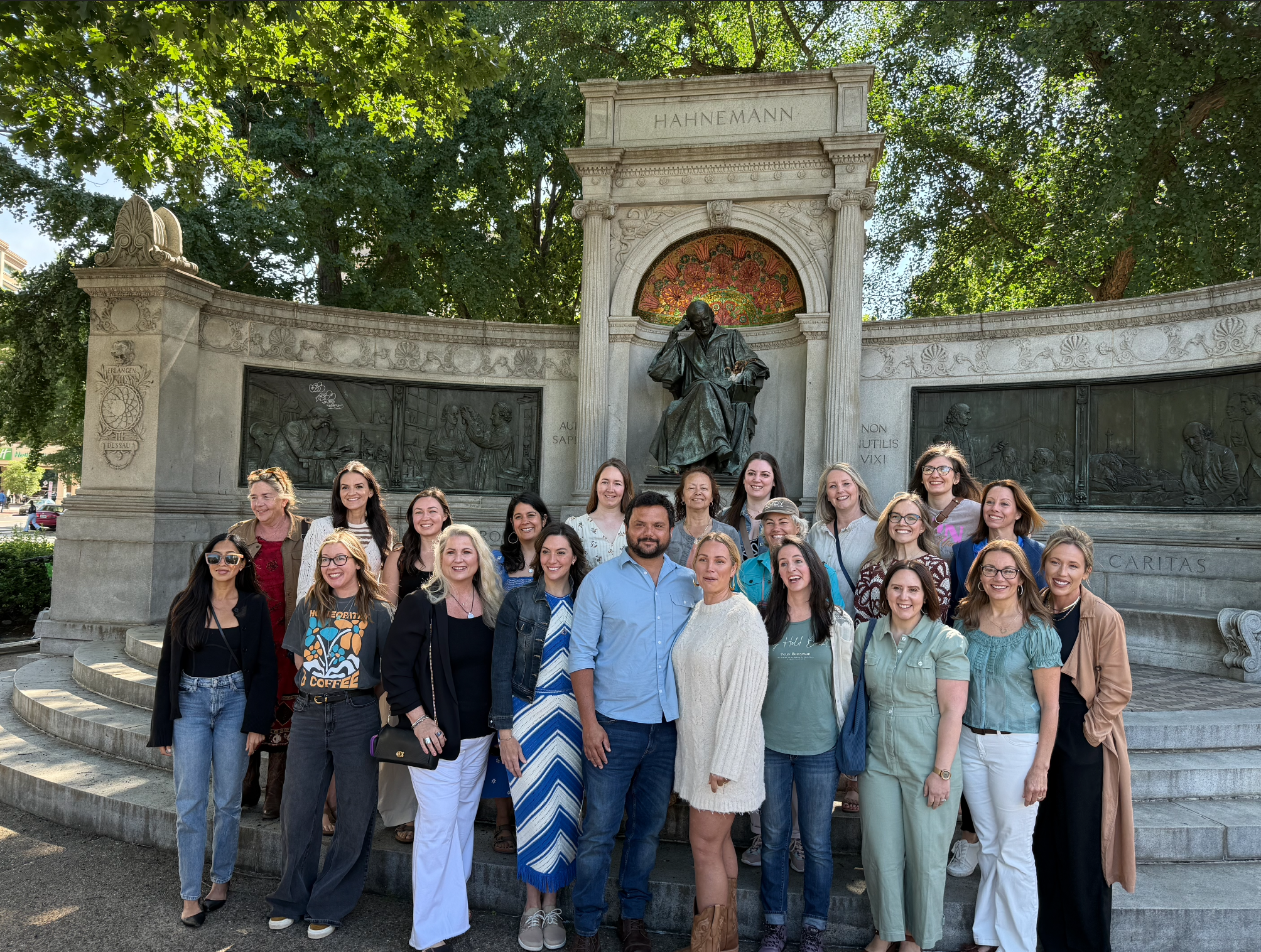 A group of diverse people smiling and standing together outdoors in front of a historic monument with sculptures and detailed carvings, surrounded by green trees.