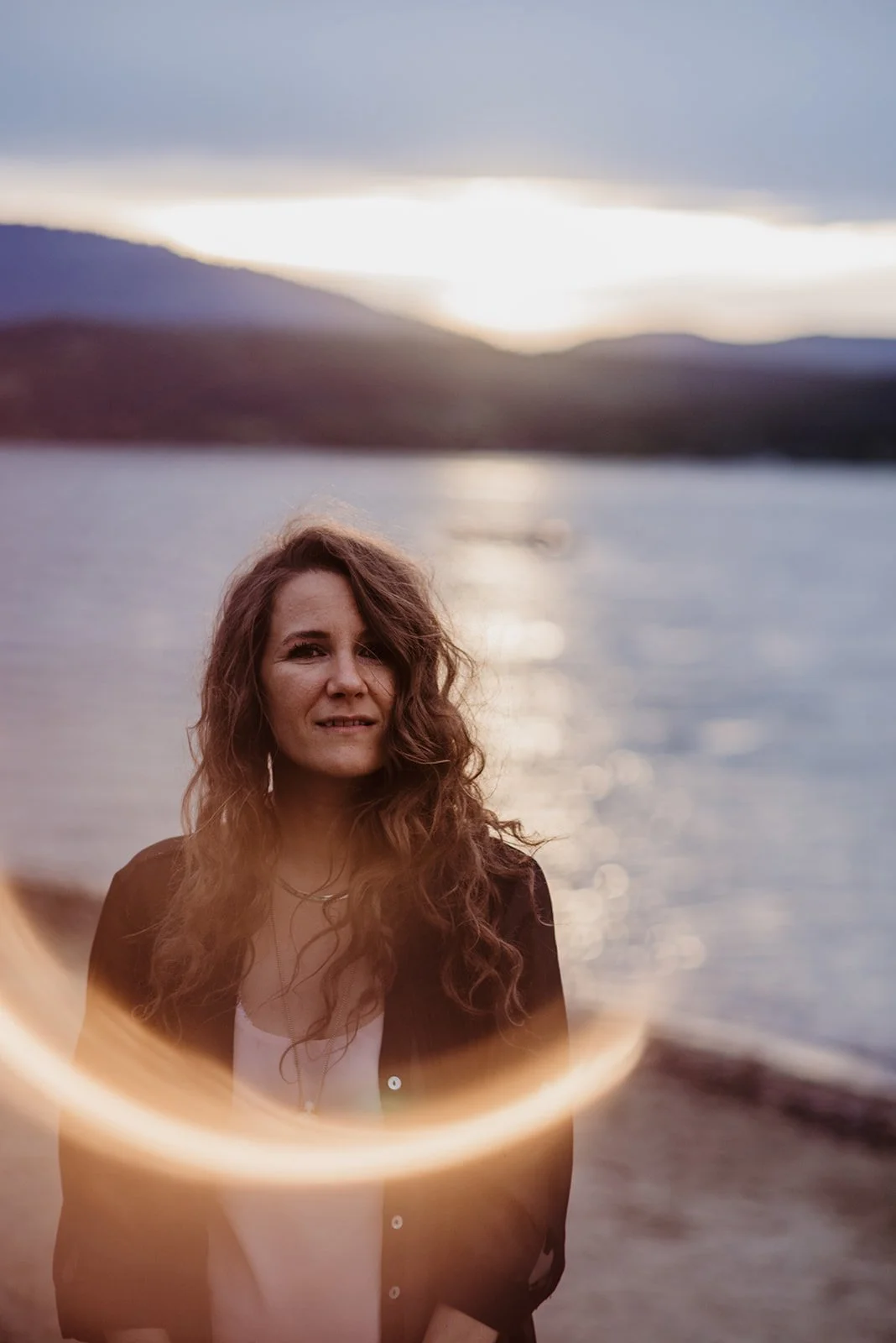 A woman with long, wavy brown hair stands in front of a body of water with mountains in the background during sunset. There is a circular light flare in the foreground.