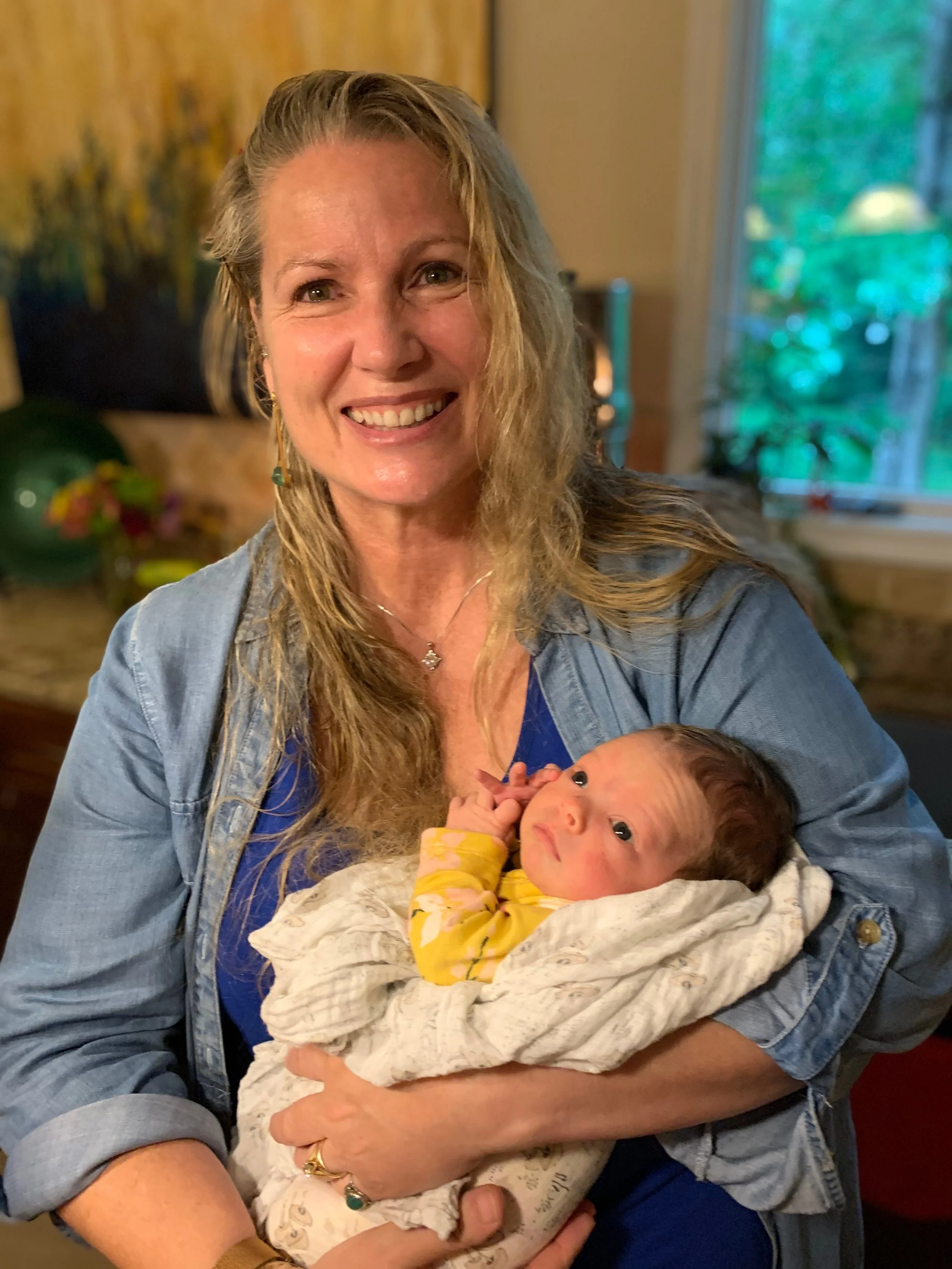 A woman smiling while holding a newborn baby wrapped in a blanket, indoors with a window showing green trees in the background.