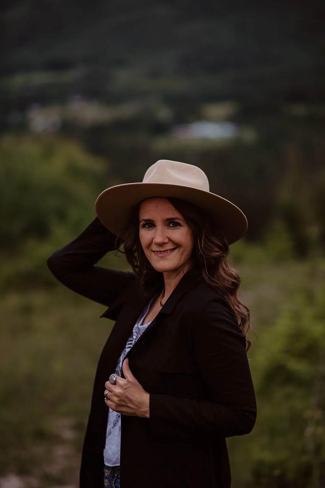 Melissa E Earle aka Mee A woman smiling, wearing a tan wide-brim hat, black jacket, and patterned shirt, standing outdoors in a green natural setting with blurred background.