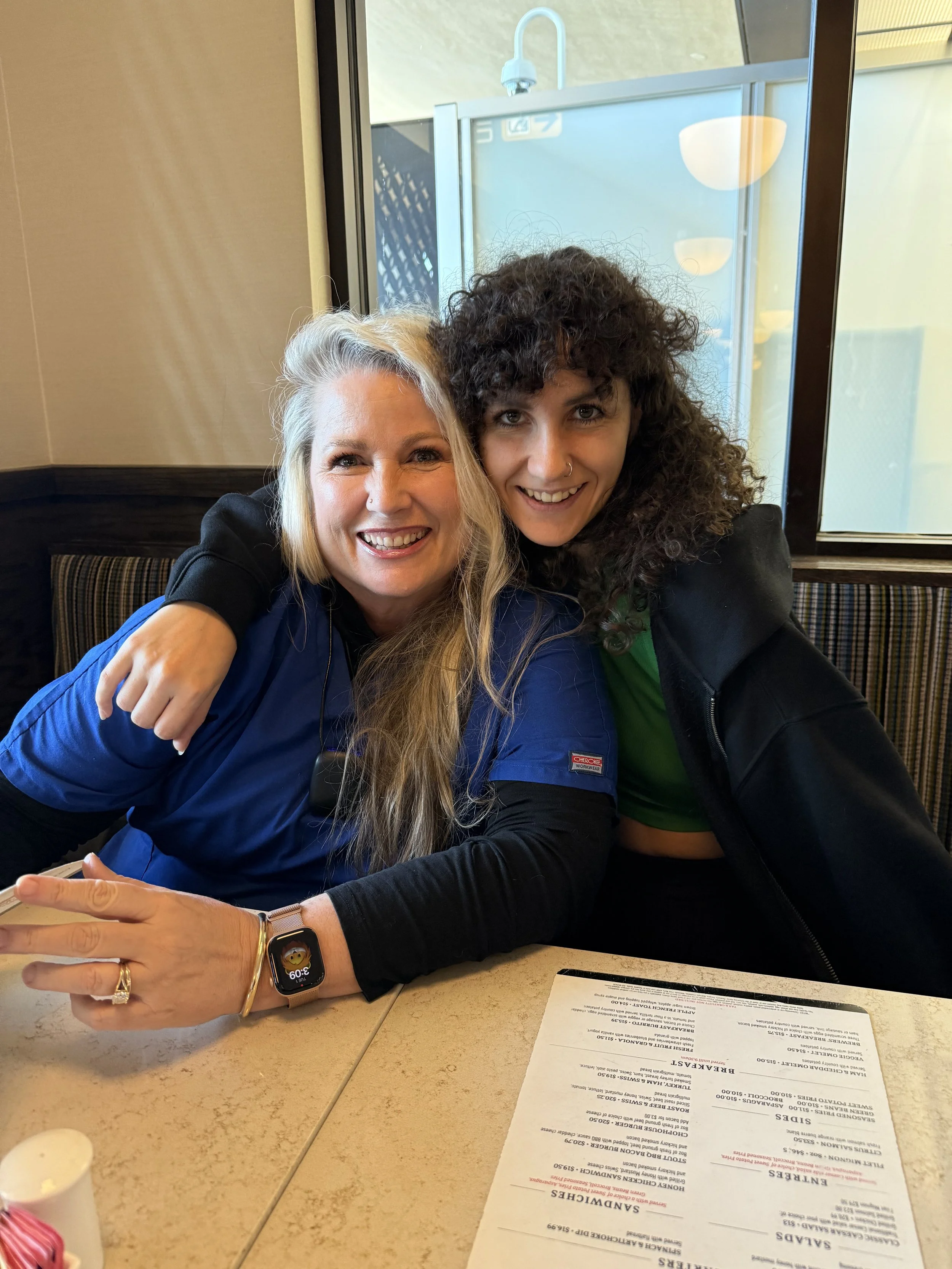 Two women are sitting together inside a restaurant, smiling and leaning on each other. One has curly dark hair and a nose piercing; the other has long blonde hair. They are at a table with a menu and a smartwatch is visible on one woman’s wrist.