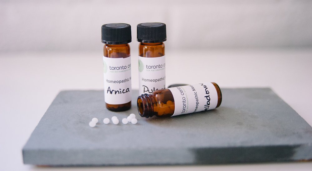 Three small bottles labeled 'Homeopathic' on a gray platform with white pills scattered nearby.