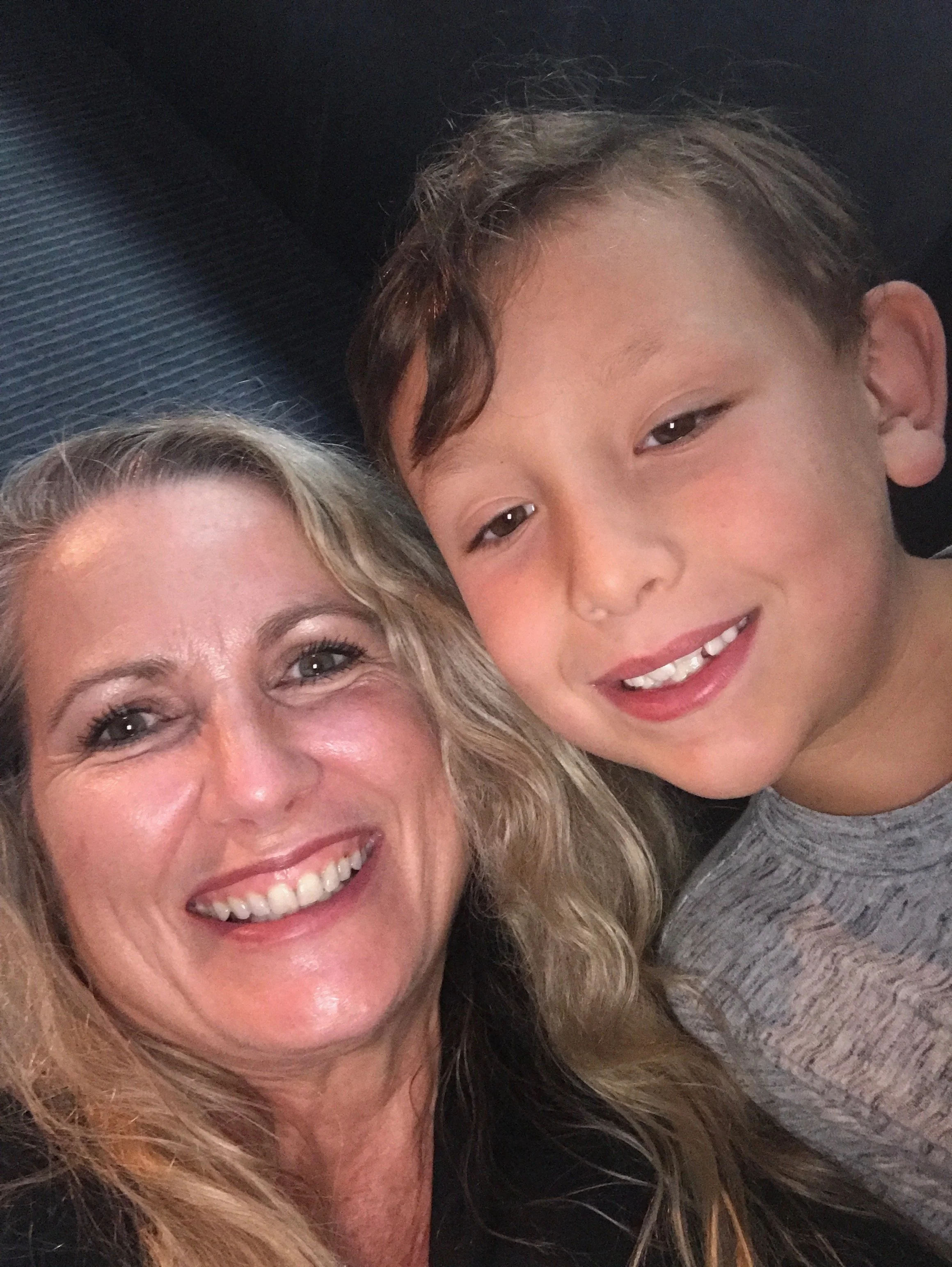 A close-up of a smiling woman with blonde, wavy hair and a young boy with short, curly hair and a wide smile, both lying on a dark textured surface.