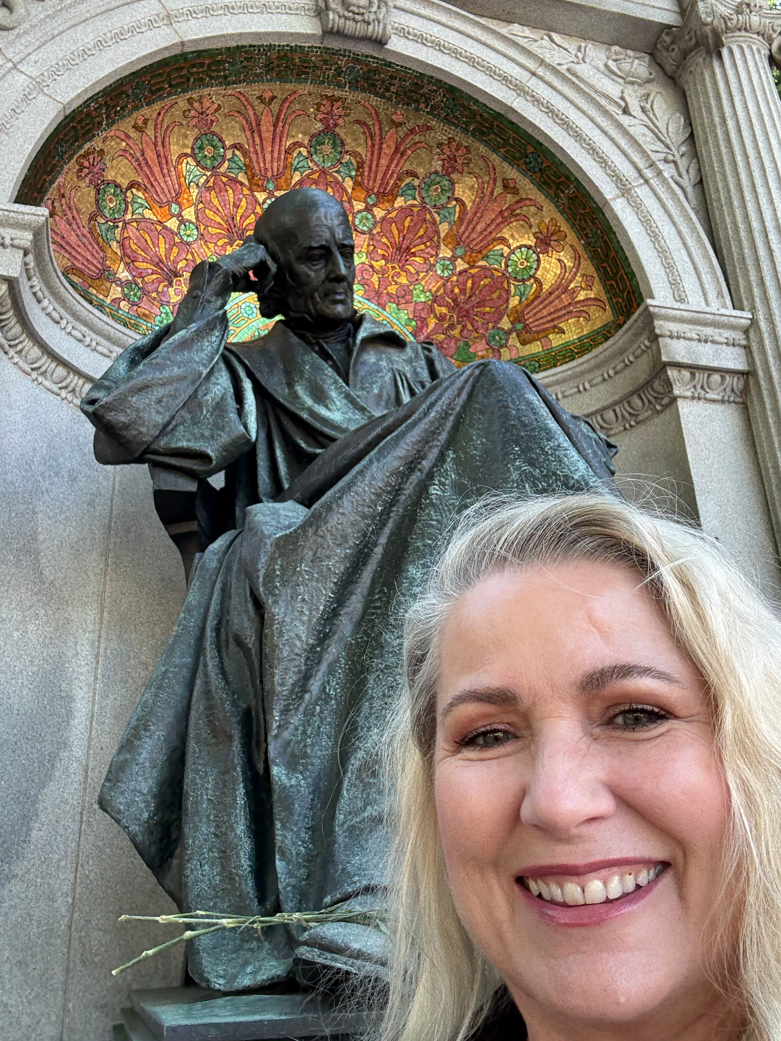 A woman with blonde hair taking a selfie in front of a statue of William Faulkner, with a decorative stained glass background featuring flowers and leaves.