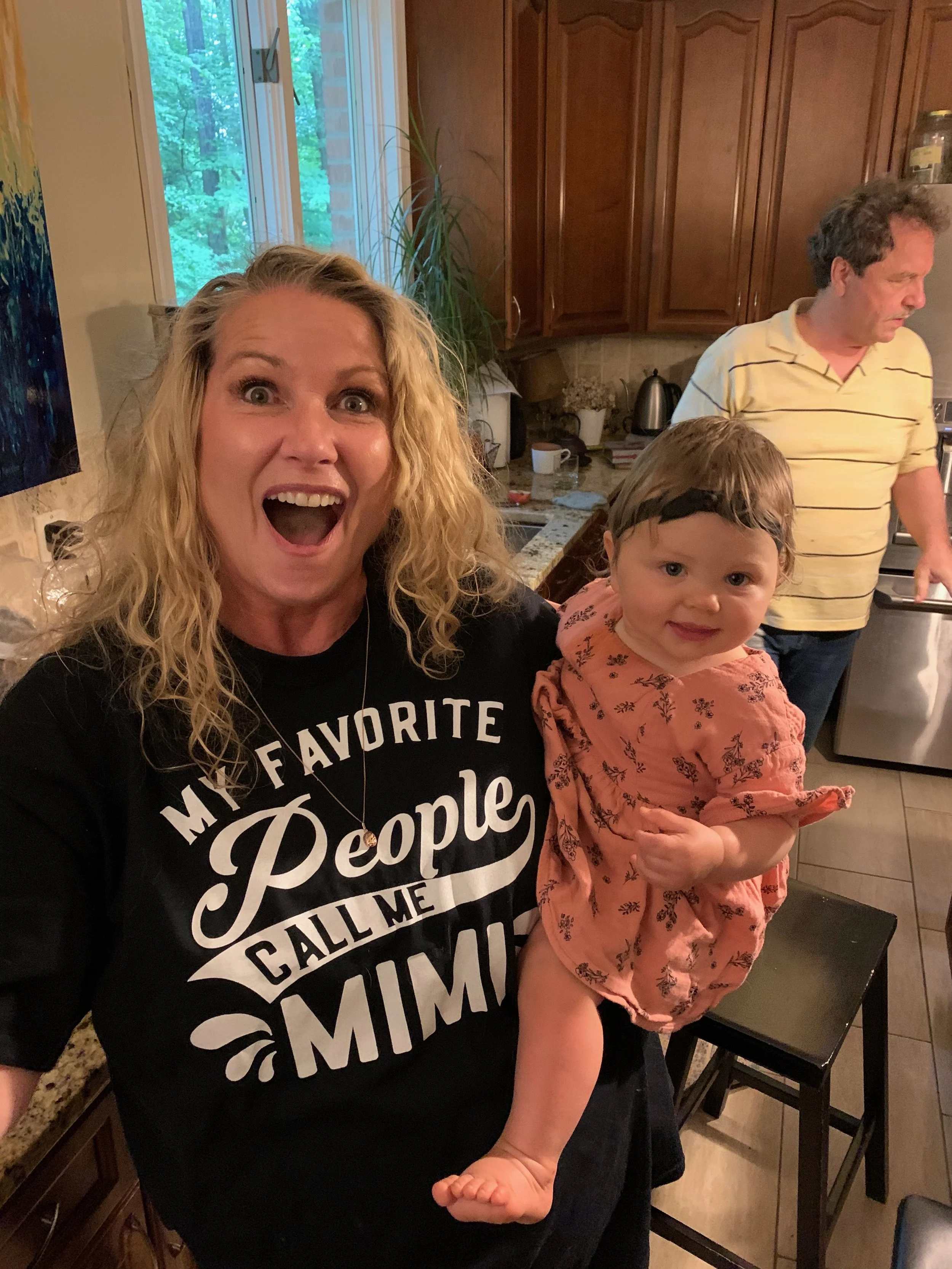 A woman with curly blonde hair taking a selfie in a kitchen, holding a young girl with curly brown hair and blue eyes. In the background, a man in a yellow striped shirt is standing near a counter. The kitchen has wooden cabinets, a window showing gr