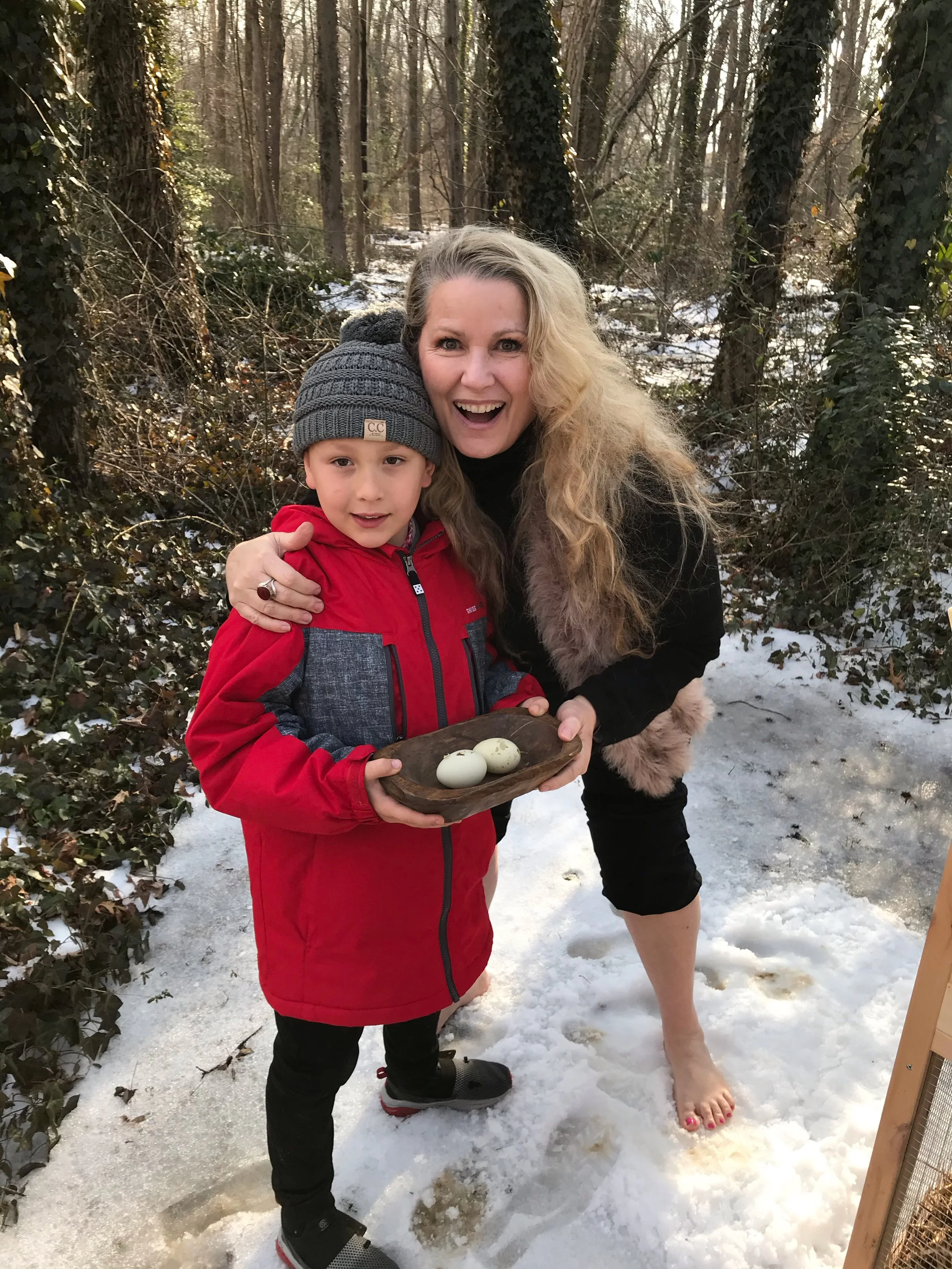 A woman with long blonde hair and a child holding a small wooden tray with three eggs, standing outdoors in a snowy wooded area. The woman is smiling and barefoot, while the child is wearing a gray beanie and red jacket.