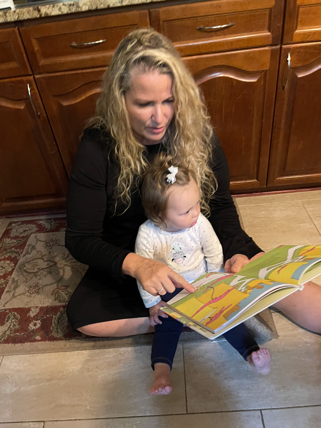 Woman and young girl sitting on the floor, reading a colorful children's book together, with wooden cabinets in the background.