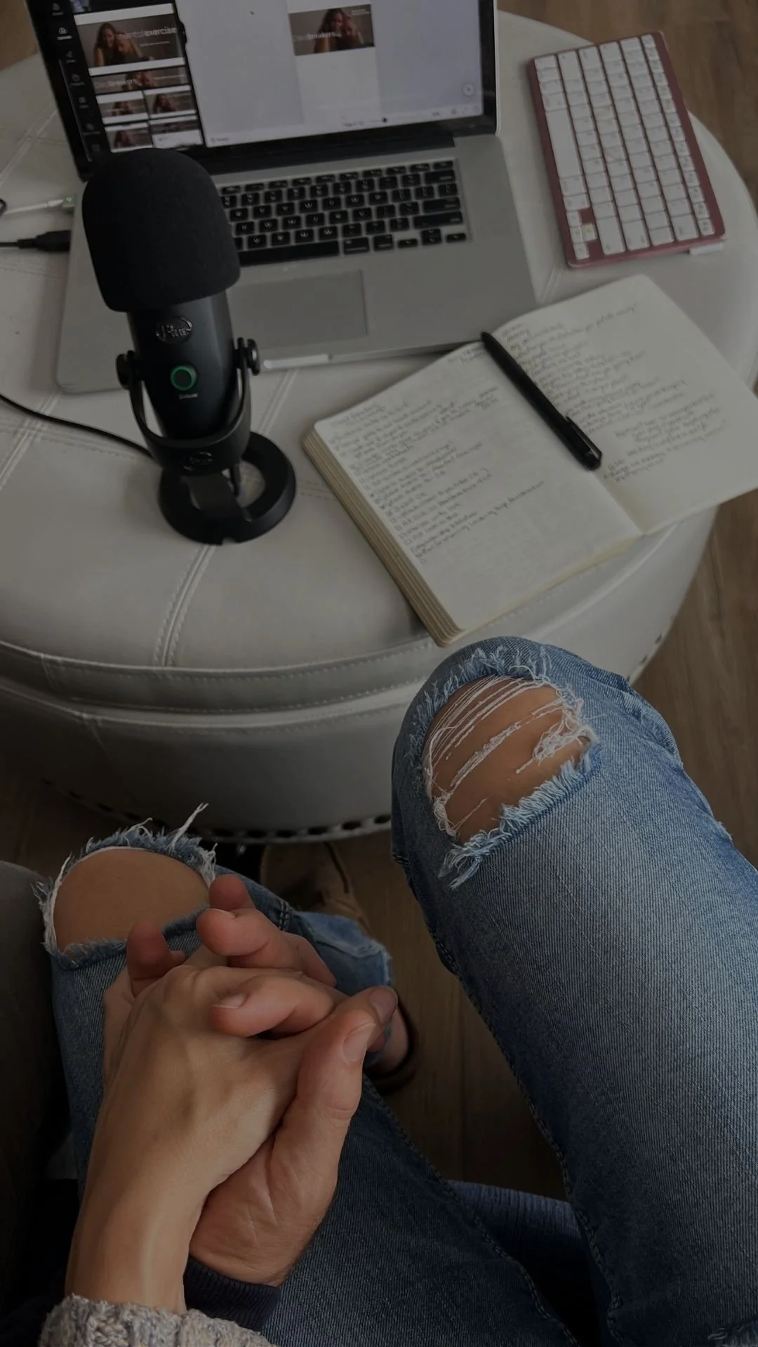 Person sitting with crossed hands, wearing ripped jeans. On the table in front, there is a laptop, a microphone, an open notebook with handwritten notes, a pen, a keyboard, and a printed sheet with images of a woman.