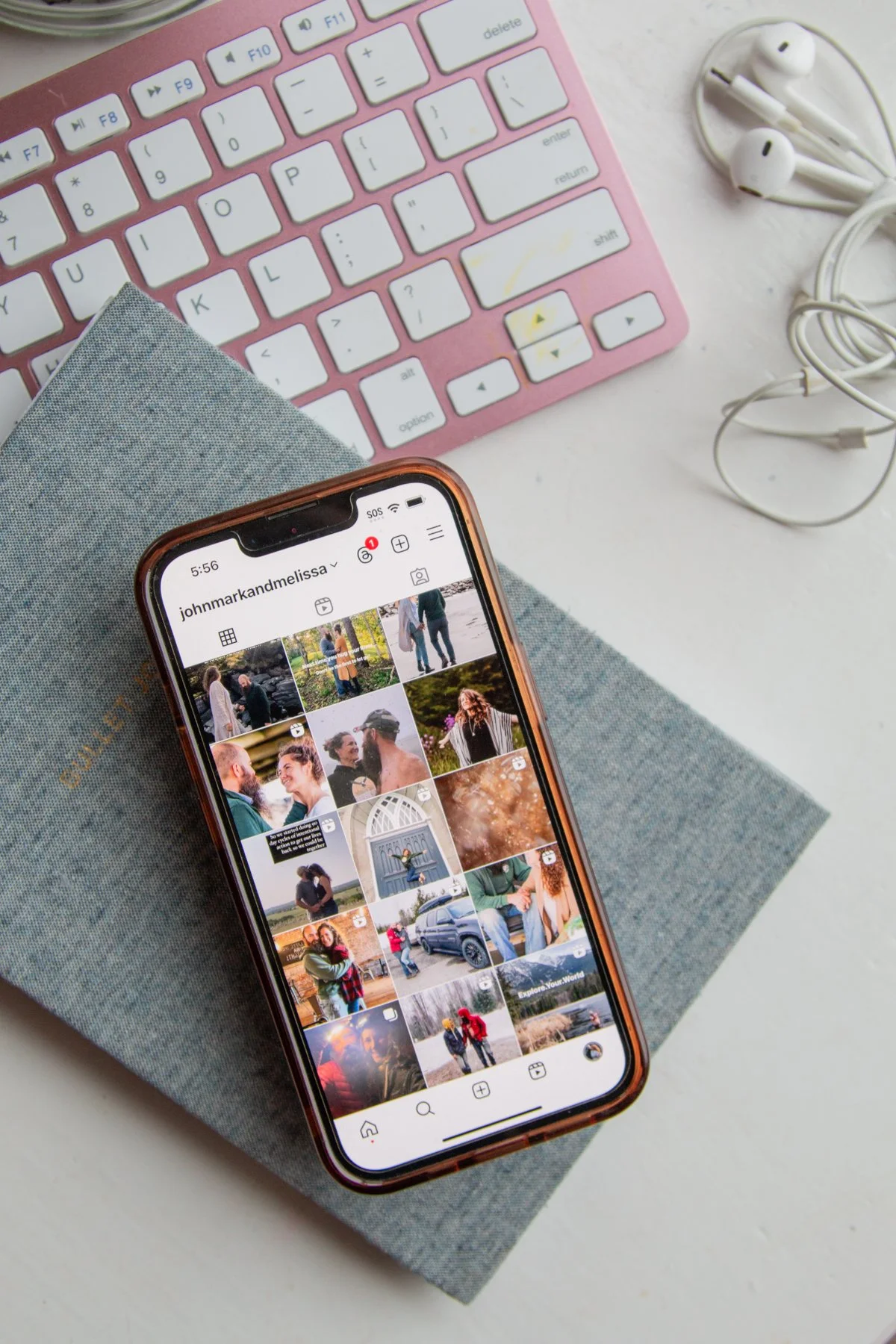 An iPhone displaying an Instagram profile page with photos of people and outdoor scenes. On a white table, there is a pink keyboard, white earbuds, and a gray fabric-covered notebook.