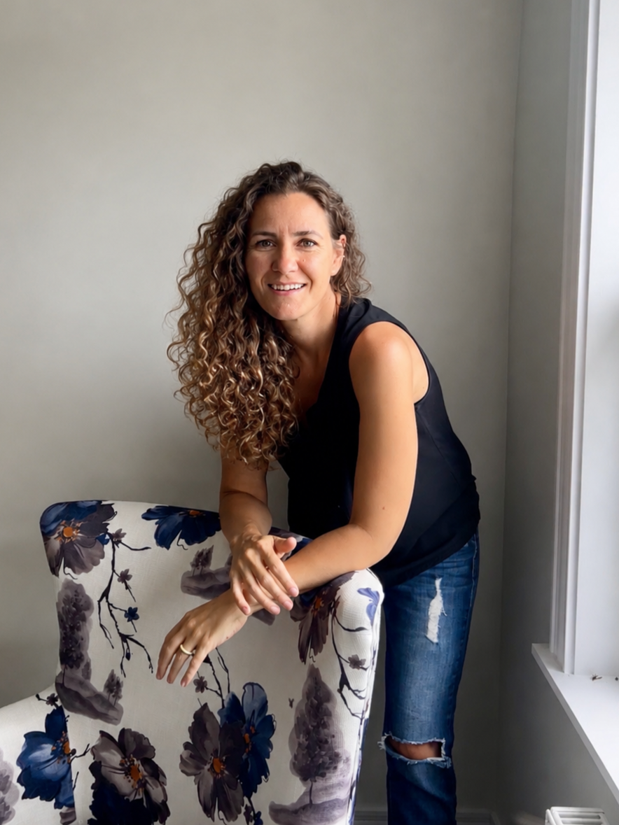 A woman with curly brown hair, wearing a black sleeveless top and ripped jeans, leaning on a floral armchair in a room with light-colored walls and a window.