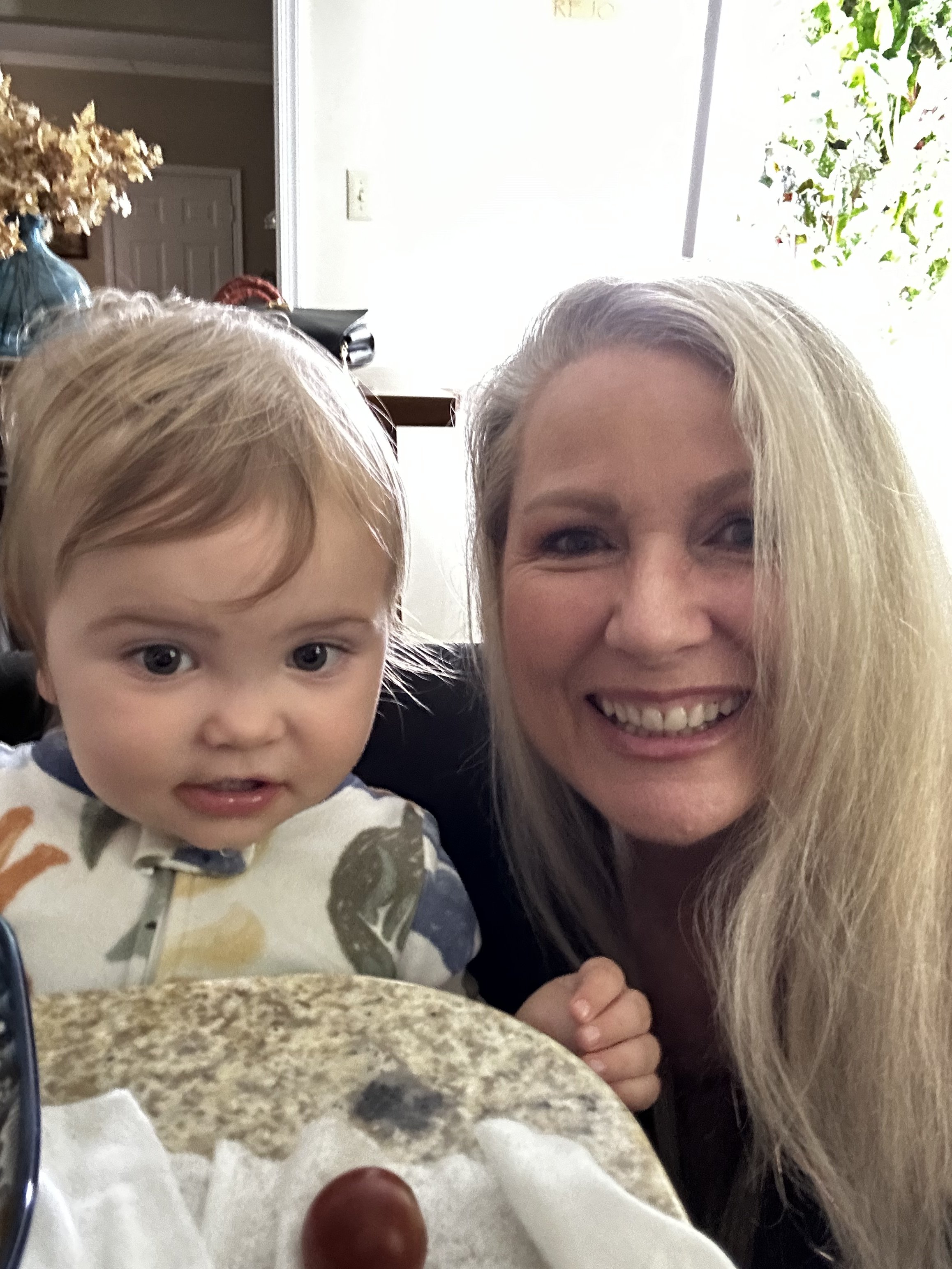 A woman with long blonde hair smiling next to a young boy with light brown hair and big eyes, both taking a selfie at a kitchen table. The table has a few grapes on it, and there is a large window with green plants in the background.