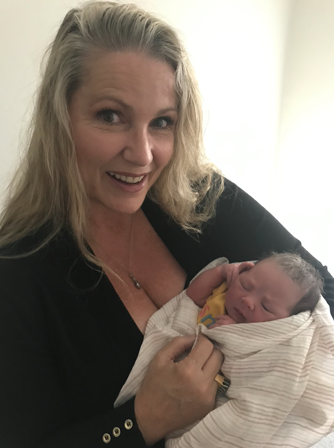 A woman with blonde hair smiling while holding a newborn baby wrapped in a striped blanket.