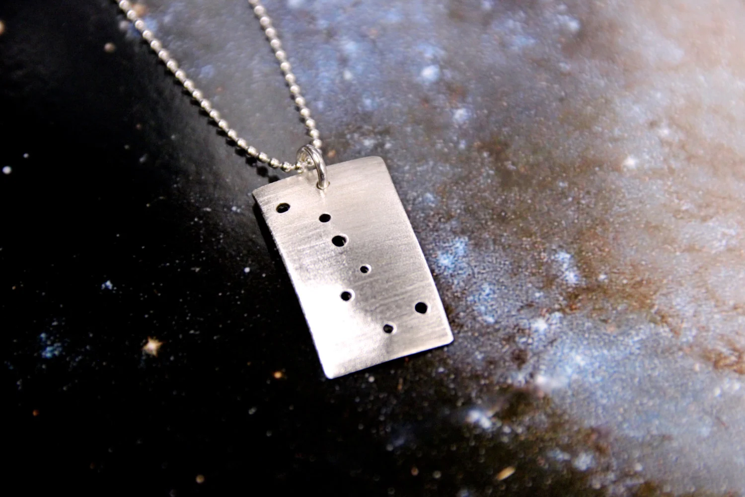 Rectangular brushed silver pendant has 7 small holes drilled to match the Big Dipper constellation. Pendant is slightly domed. Rectangle is in a vertical orientation.