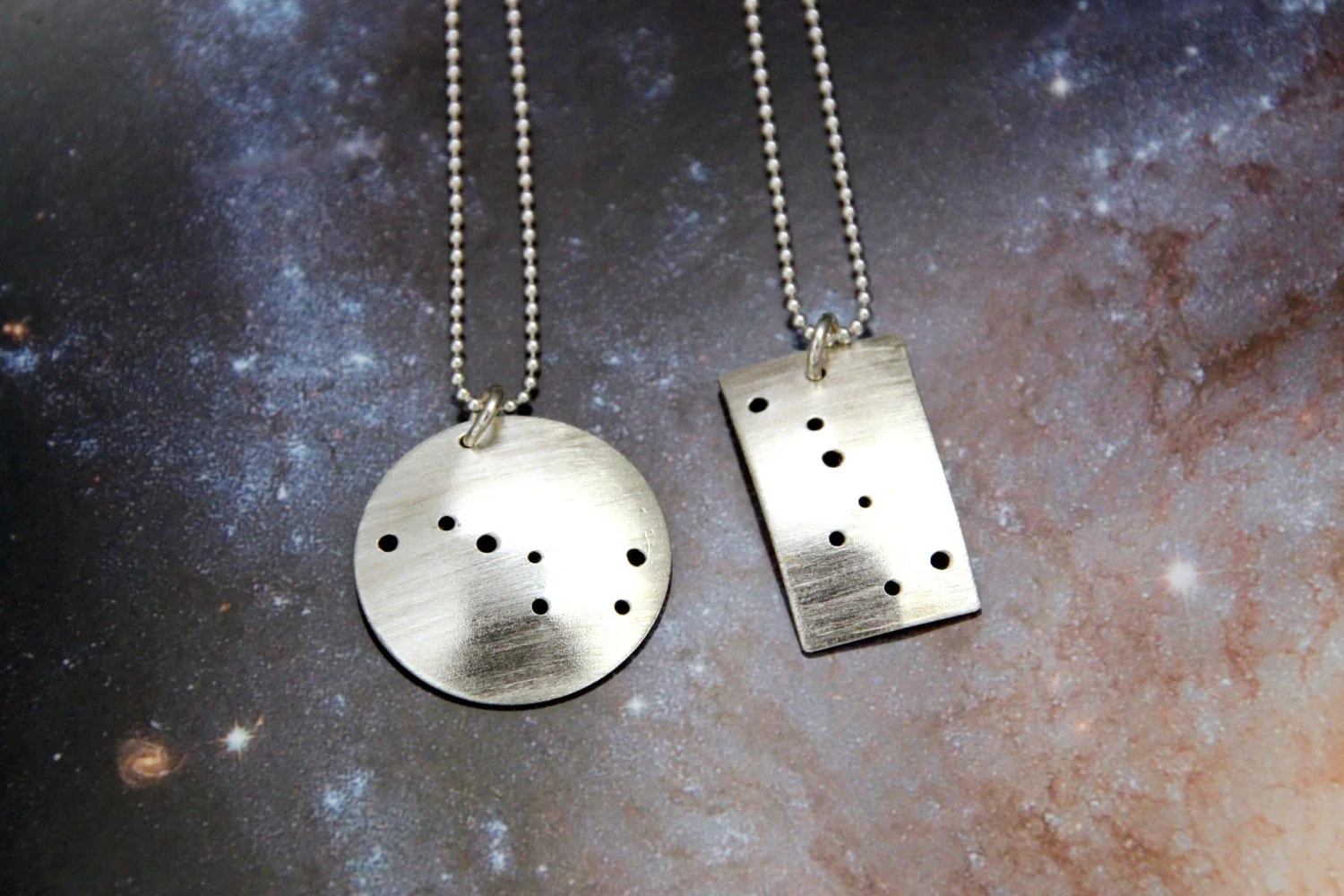 One round and one rectangular brushed silver pendant each has 7 small holes drilled to match the Big Dipper constellation. Pendants are slightly domed. Rectangle oriented vertically.