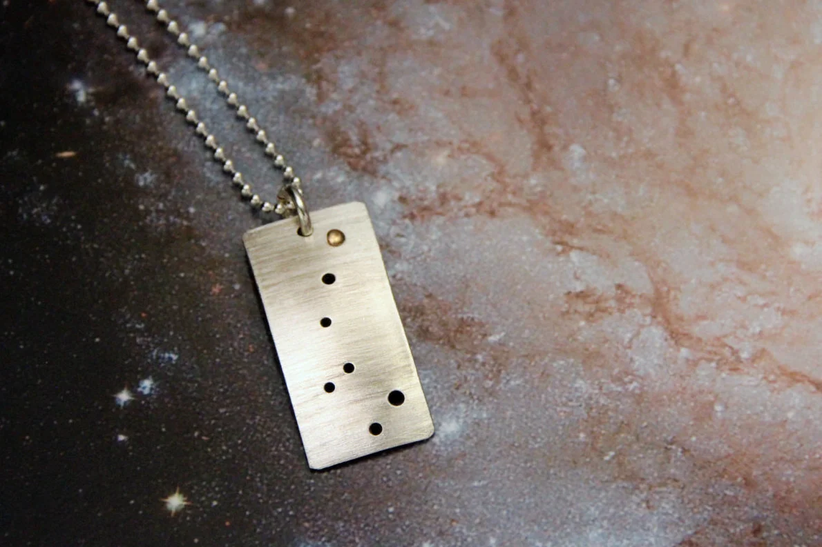 Rectangular brushed silver pendant has 7 small holes drilled to match the Little Dipper. Gold accent on the top hole represents the North Star. Pendant is slightly domed and hangs vertically.