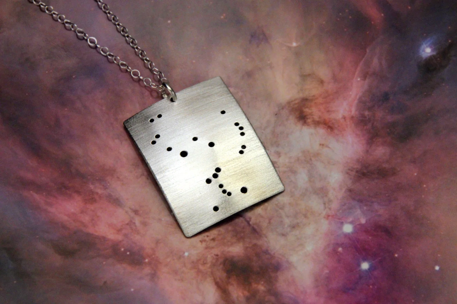 Rectangular brushed silver pendant has 22 small holes drilled to match the Orion constellation. Pendant is slightly domed. Rectangle is in a vertical orientation.