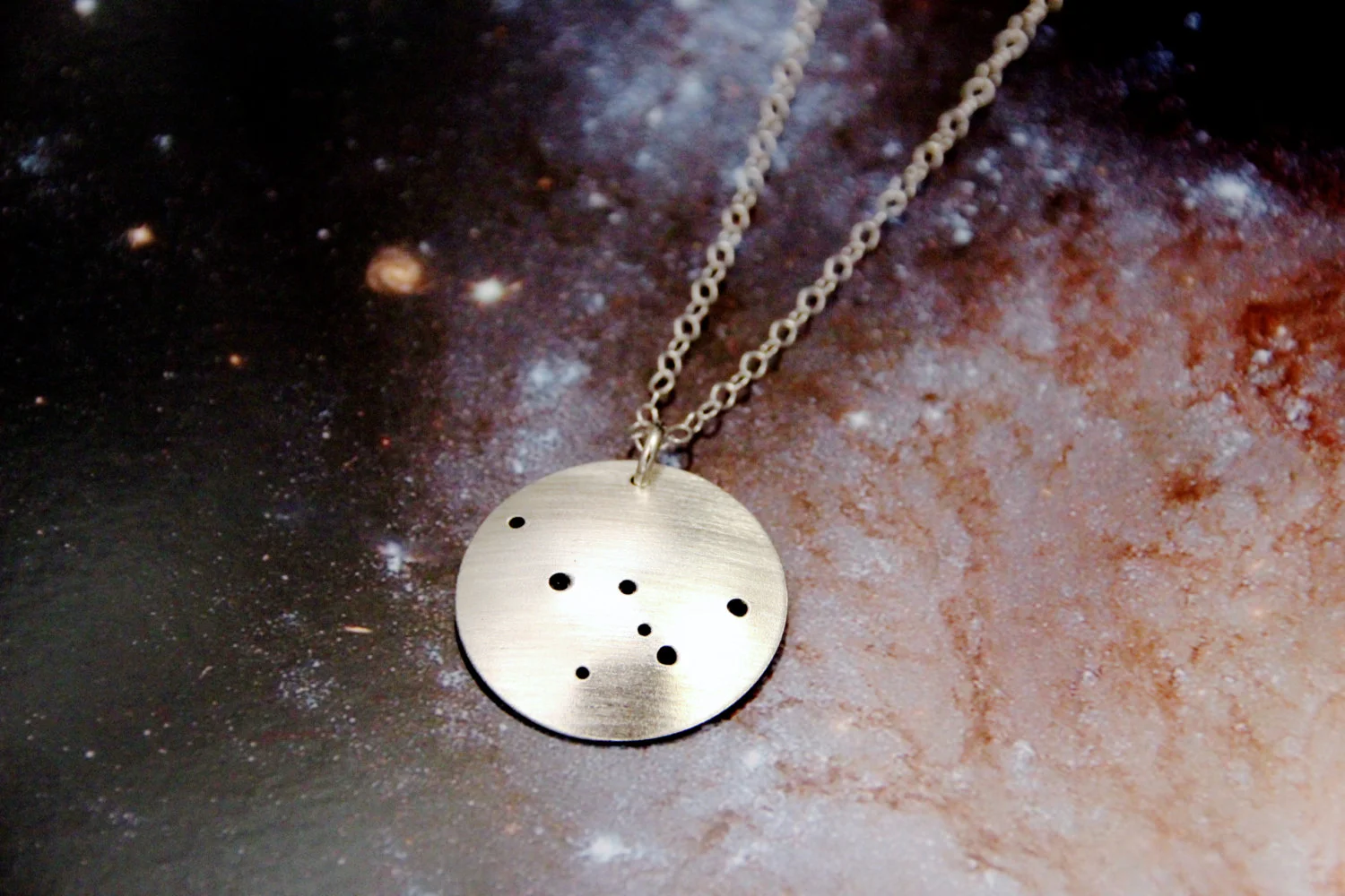 Round brushed silver pendant has 7 small holes drilled to match the Cassiopeia constellation. Pendant is slightly domed and attached to an open-link silver chain with a silver jump ring.