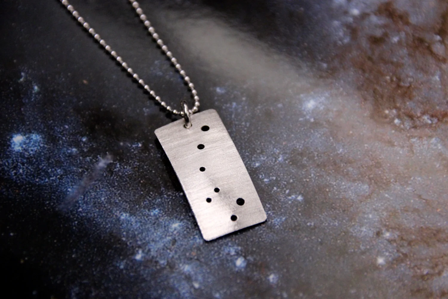 Rectangular brushed silver pendant has 7 small holes drilled to match the Little Dipper constellation. Pendant is slightly domed. Rectangle is in a vertical orientation.