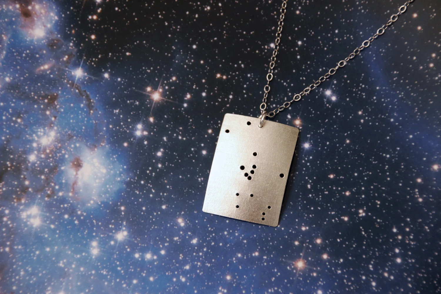 Rectangular brushed silver pendant has 15 small holes drilled to match the Taurus constellation. Pendant is slightly domed. Rectangle is in a vertical orientation.