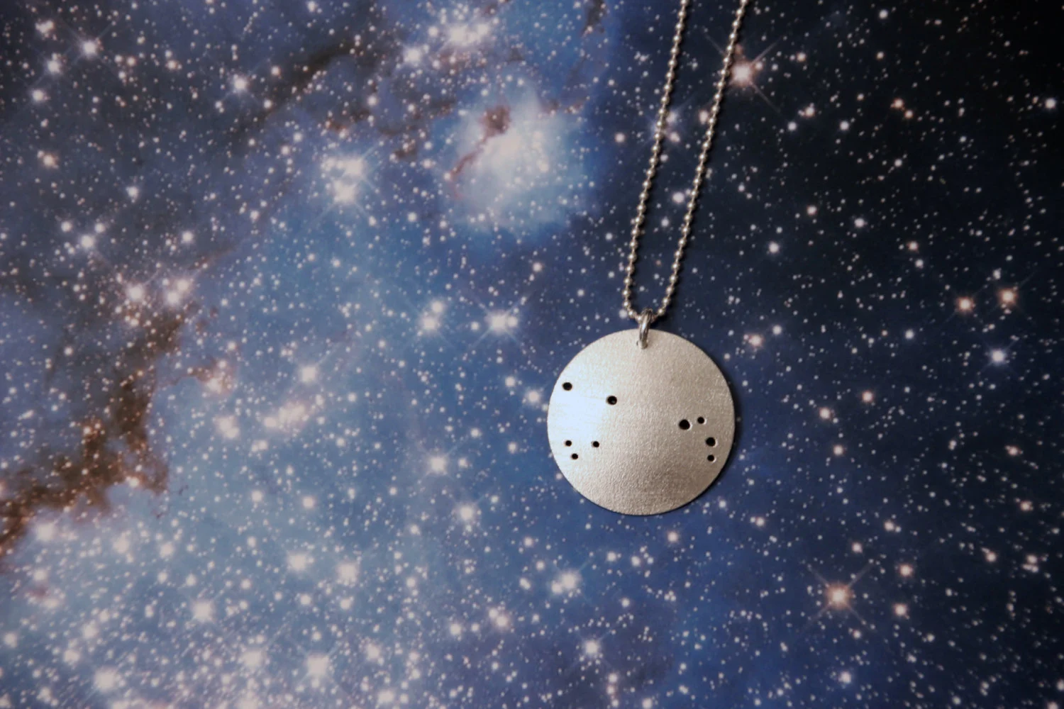 Round brushed silver pendant has 9 small holes drilled to match the Aries constellation. Pendant is slightly domed and attached to a narrow silver ball chain with a silver jump ring.