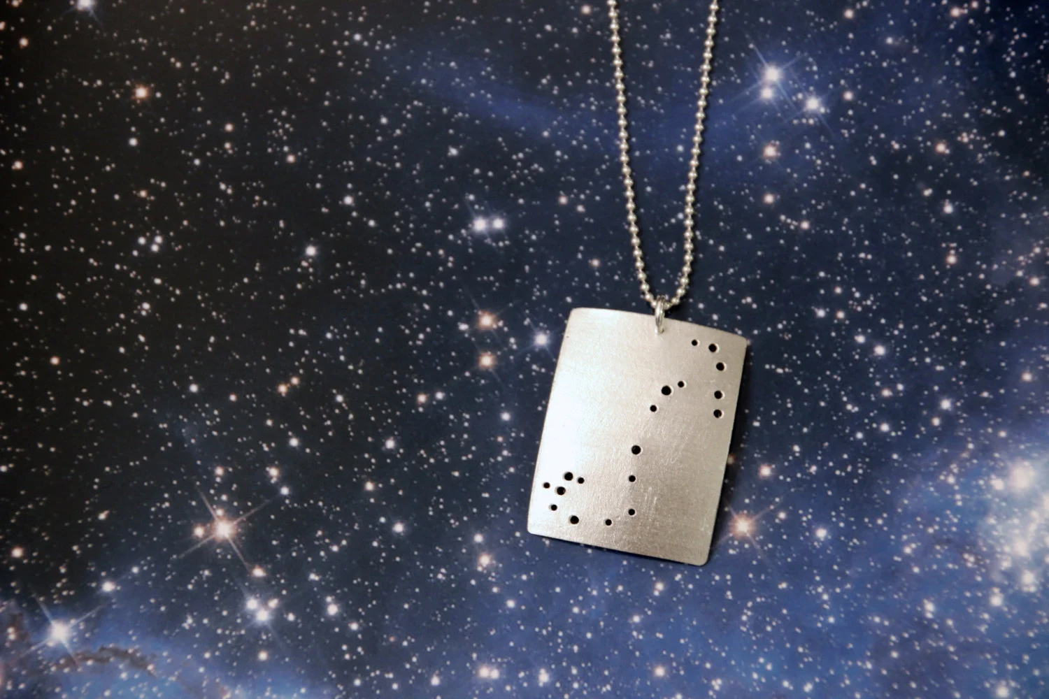 Rectangular brushed silver pendant has 18 small holes drilled to match the Scorpio constellation. Pendant is slightly domed. Rectangle is in a vertical orientation.