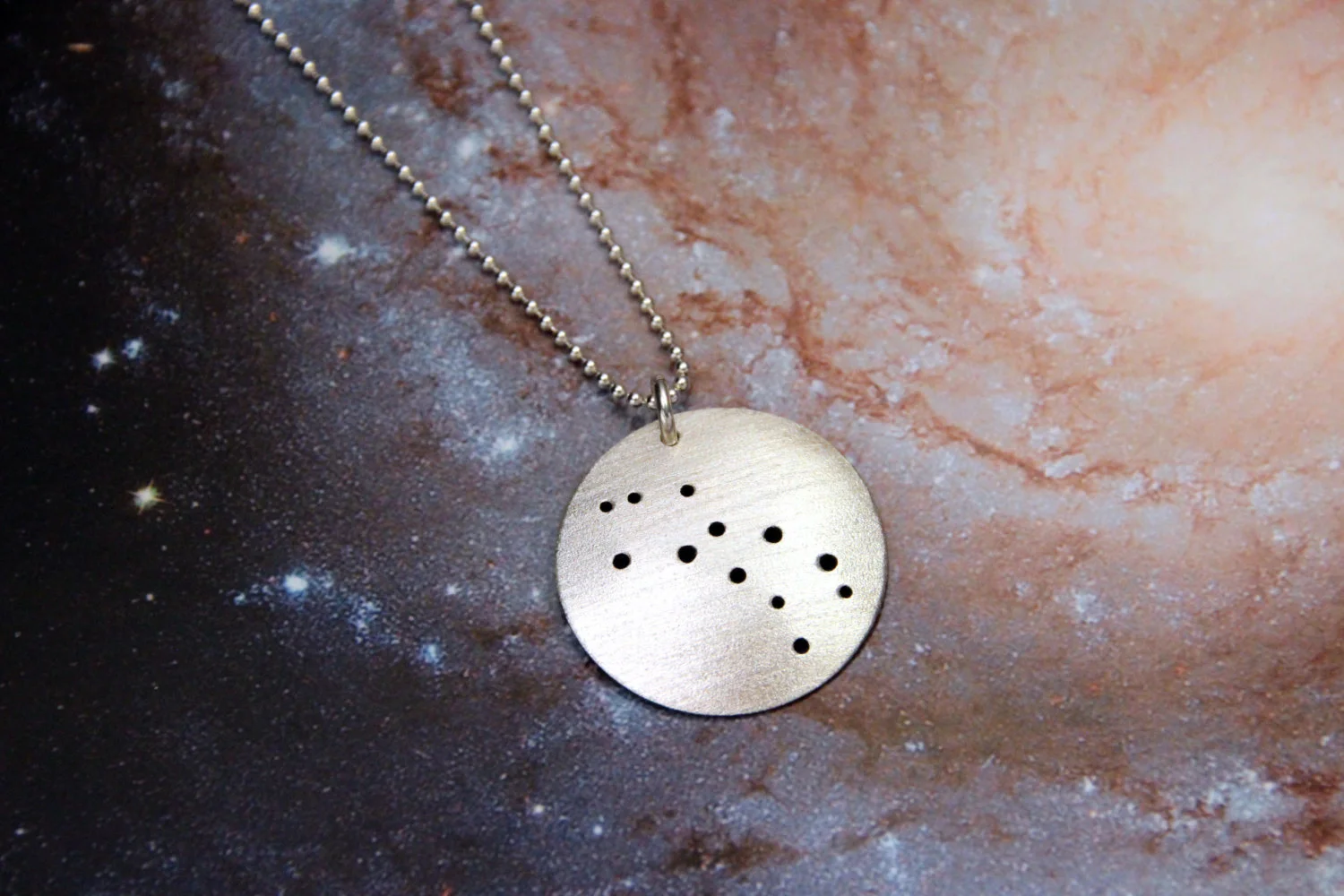 Round brushed silver pendant has 12 small holes drilled to match the Lupus constellation. Pendant is slightly domed and attached to a narrow silver ball chain with a silver jump ring.