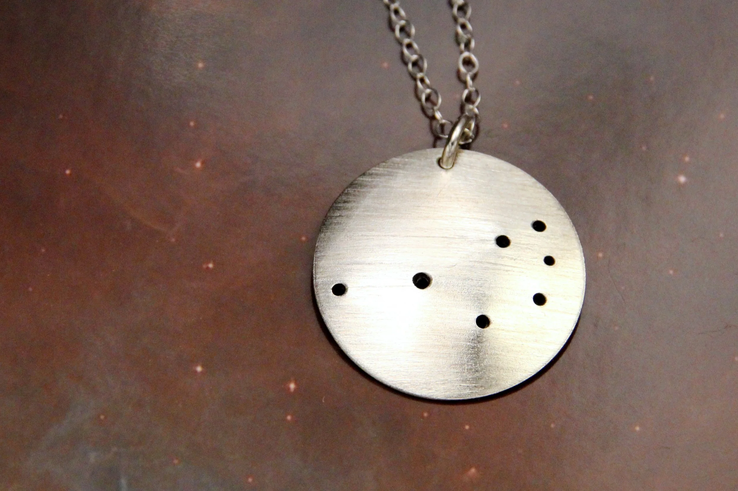 Round brushed silver pendant has 7 small holes drilled to match the Pleiades constellation. Pendant is slightly domed and attached to an open-link silver chain with a silver jump ring.
