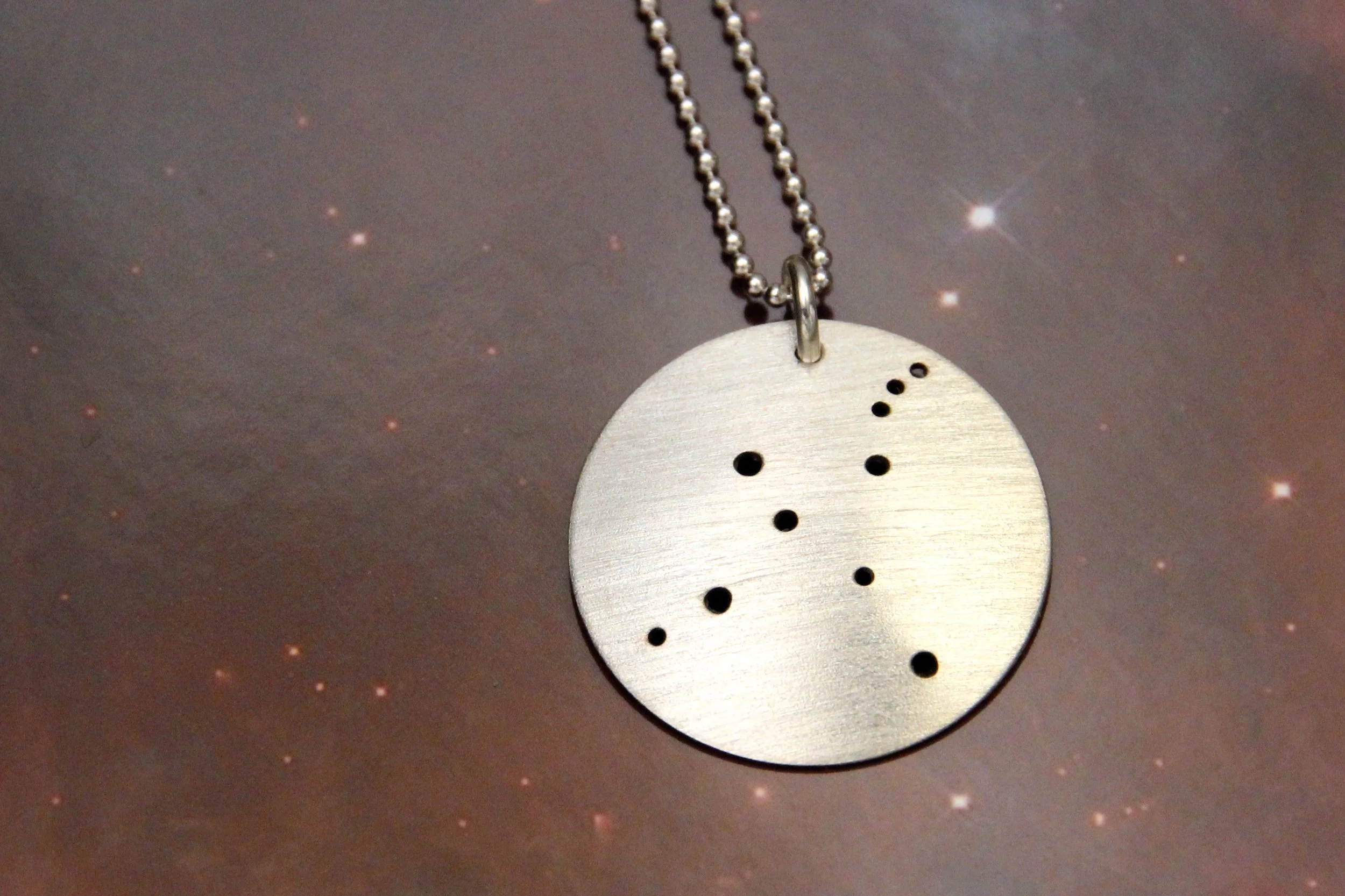 Round brushed silver pendant has 10 small holes drilled to match the Cygnus constellation. Pendant is slightly domed and attached to a narrow silver ball chain with a silver jump ring.