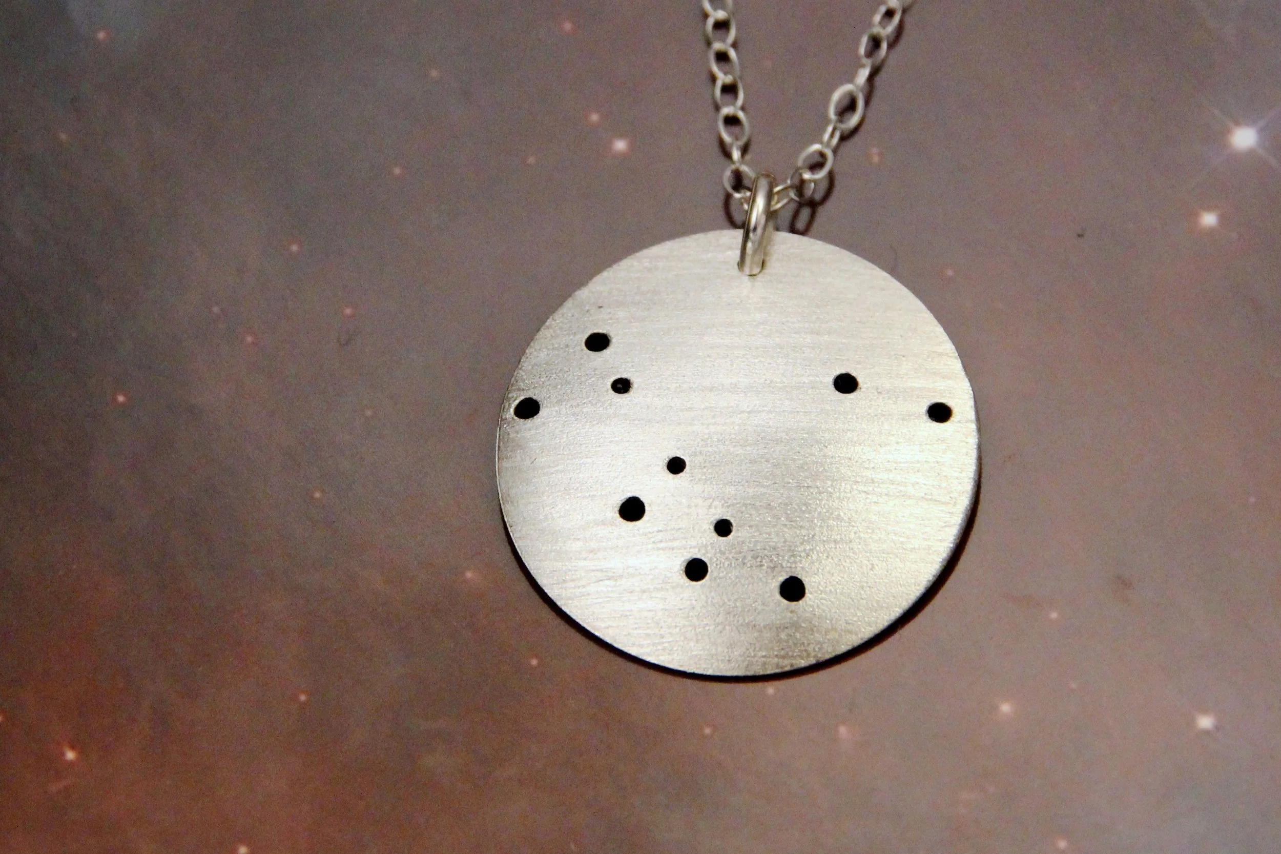 Round brushed silver pendant has 10 small holes drilled to match the Andromeda constellation. Pendant is slightly domed and attached to an open-link silver chain with a silver jump ring.