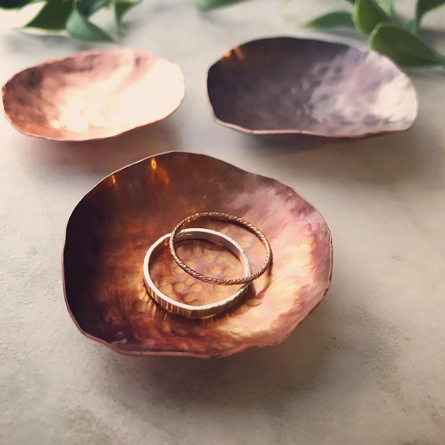 3 small copper ring dishes are shown up close. In the front is colorful orange-pink with 2 plain rings inside to show the 2” scale. Behind are bright raw copper and darkened brownish-purple dishes.