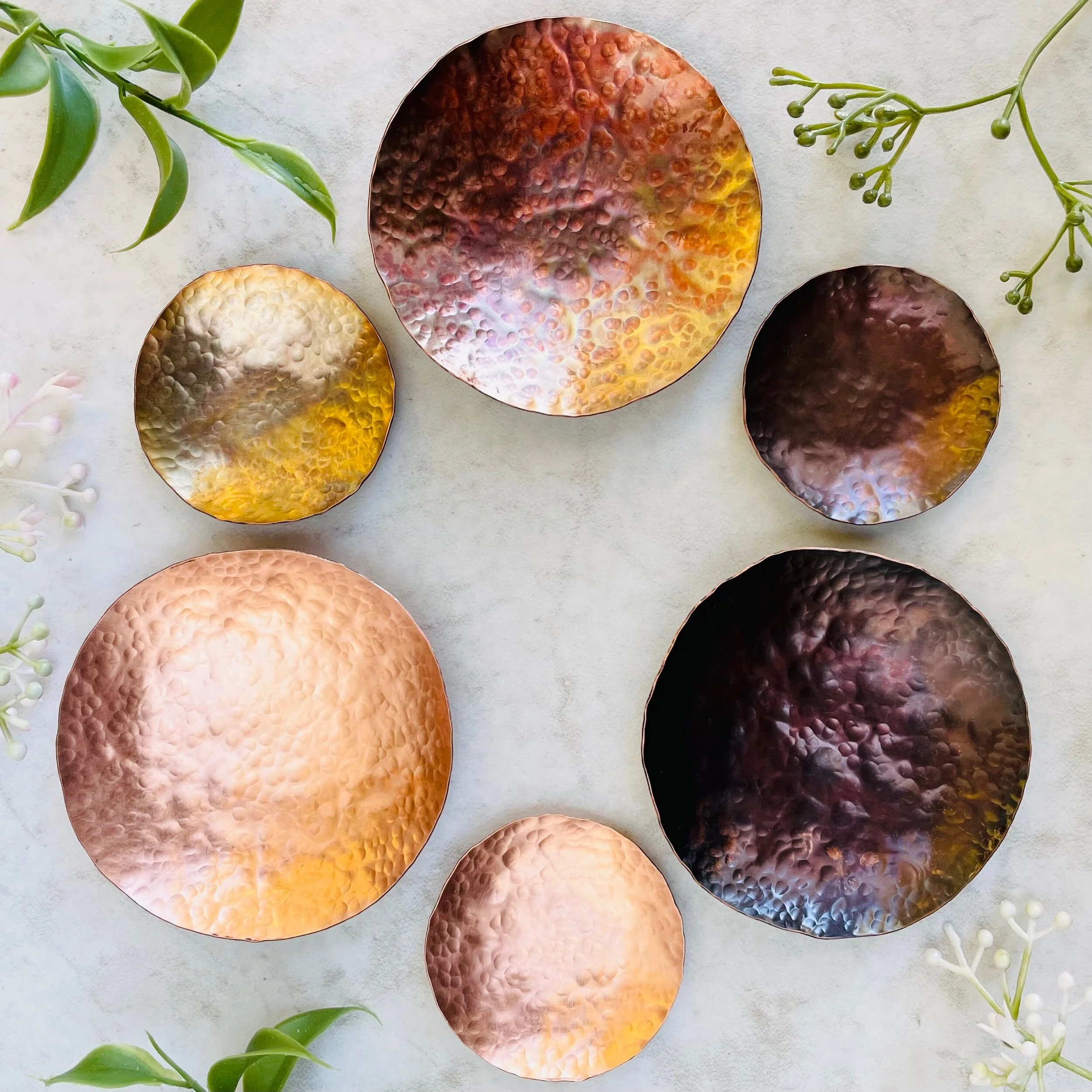 Six hammer textured copper ring dishes are arranged in a circle. The bright raw copper, assorted colorful copper, and darkened copper finishes are each shown on a small and large dish.